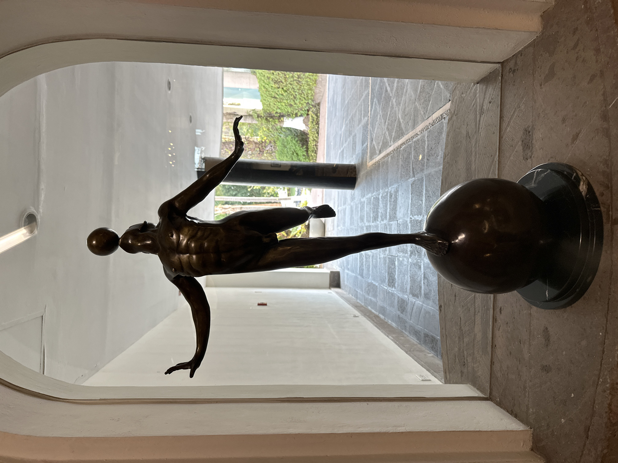 A bronze sculpture of a nude male figure balancing on a sphere is centered in an arched doorway or opening. The figure is oriented horizontally, with a sphere balanced on his head. He is captured in a dynamic pose with one arm raised and the other extended downwards, while one leg is extended and resting on the larger sphere. The sculpture rests on a round, dark-colored base.
The location appears to be an interior space leading to an exterior area, evidenced by the greenery visible through the opening. The interior walls are light-colored, and the floor is tiled. The exterior appears to be a courtyard or garden setting.
The location is identified as San Bernardino Tlaxcalancingo, Mexico.