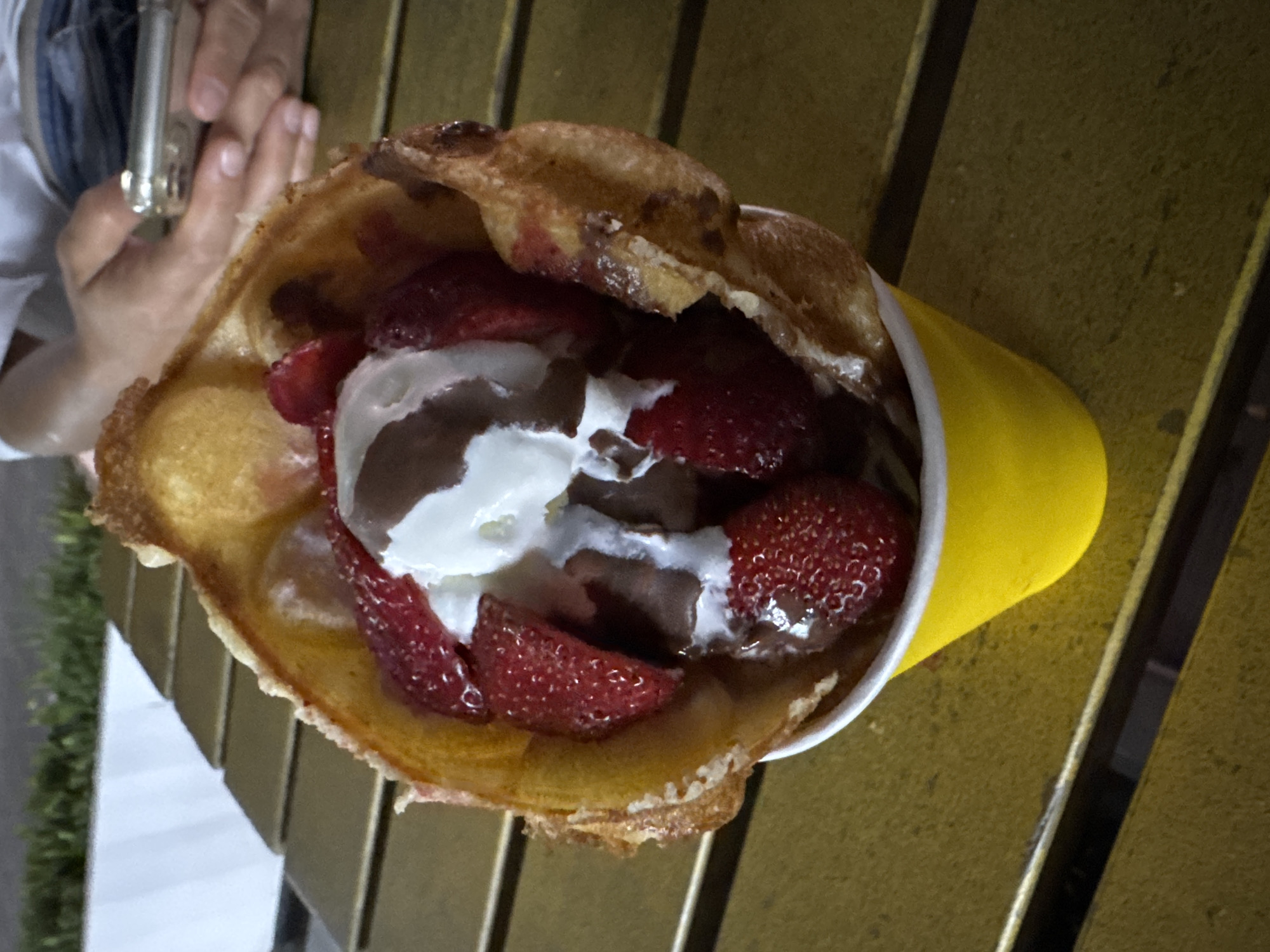 A close-up, high-angle shot shows a bubble waffle cone filled with ice cream, strawberries, and chocolate sauce. The waffle is golden brown and crispy, with a slightly irregular shape. Inside, scoops of white ice cream are interspersed with sliced strawberries and drizzles of dark chocolate. The cone sits in a bright yellow paper cup, which is placed on a textured, golden-brown surface that appears to be a wooden table. In the background, out of focus, is a person holding a mobile phone and wearing a white shirt and blue jeans. Beyond that, a blurry green area suggests vegetation and a white structure. The image appears to be taken outdoors, likely in a cafe or outdoor seating area in Zharbulak, Kazakhstan.