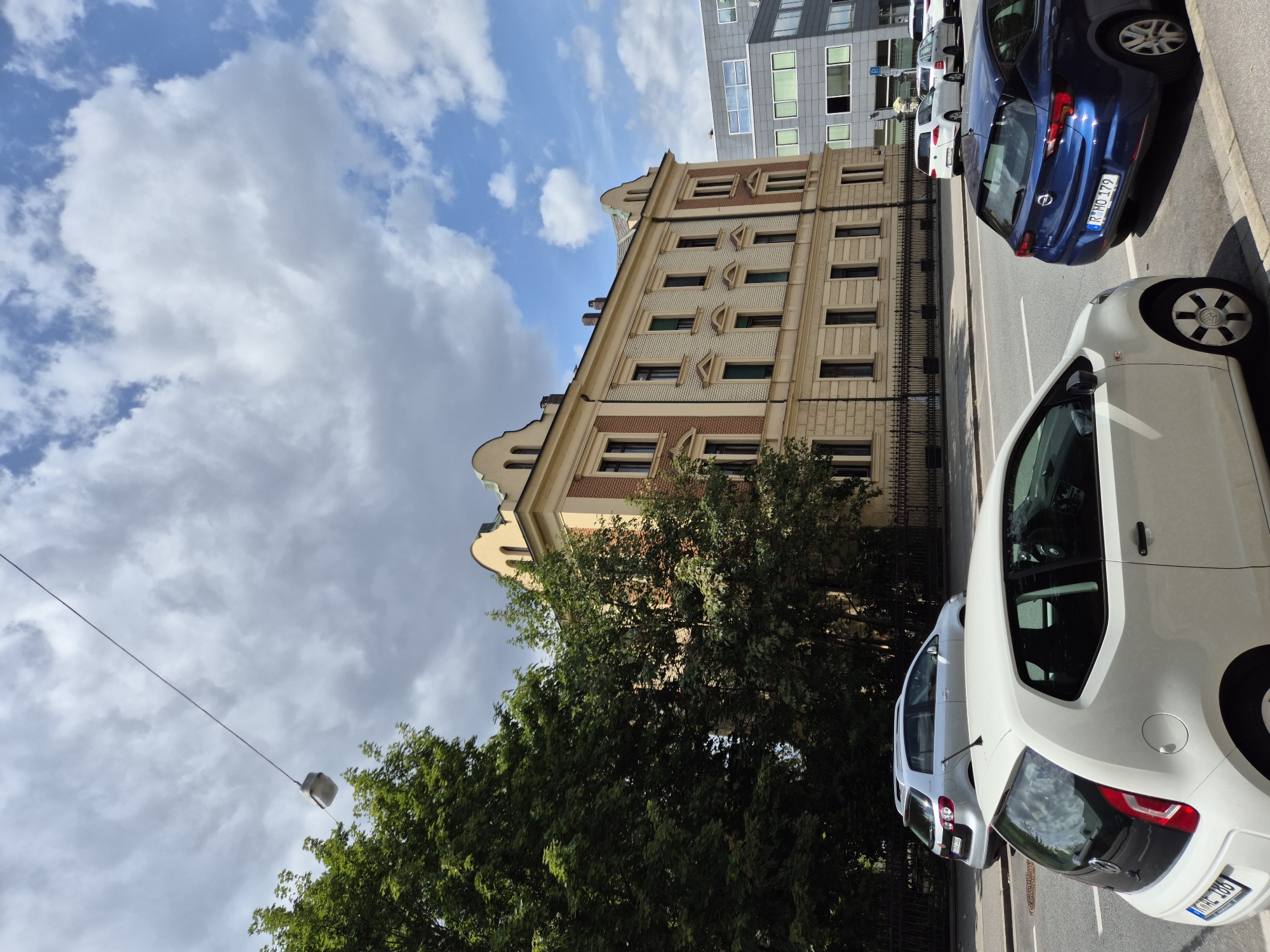 The image depicts a street scene in Regensburg, Germany, under a partly cloudy sky. In the foreground, a white Volkswagen Up! is parked on the right side of the road. To its right, a blue Opel Astra hatchback is also parked. In the middle ground, a silver Volkswagen Golf station wagon is driving on the road. Behind the cars, a historic, light yellow building with red brick accents and arched windows stands prominently. To the right of this building, a modern, gray multi-story office building with a glass facade is visible. Large trees line the left side of the street, partially obscuring the historic building. A streetlight hangs over the road from a cable. The road appears to be a typical urban street with painted lane markings.
