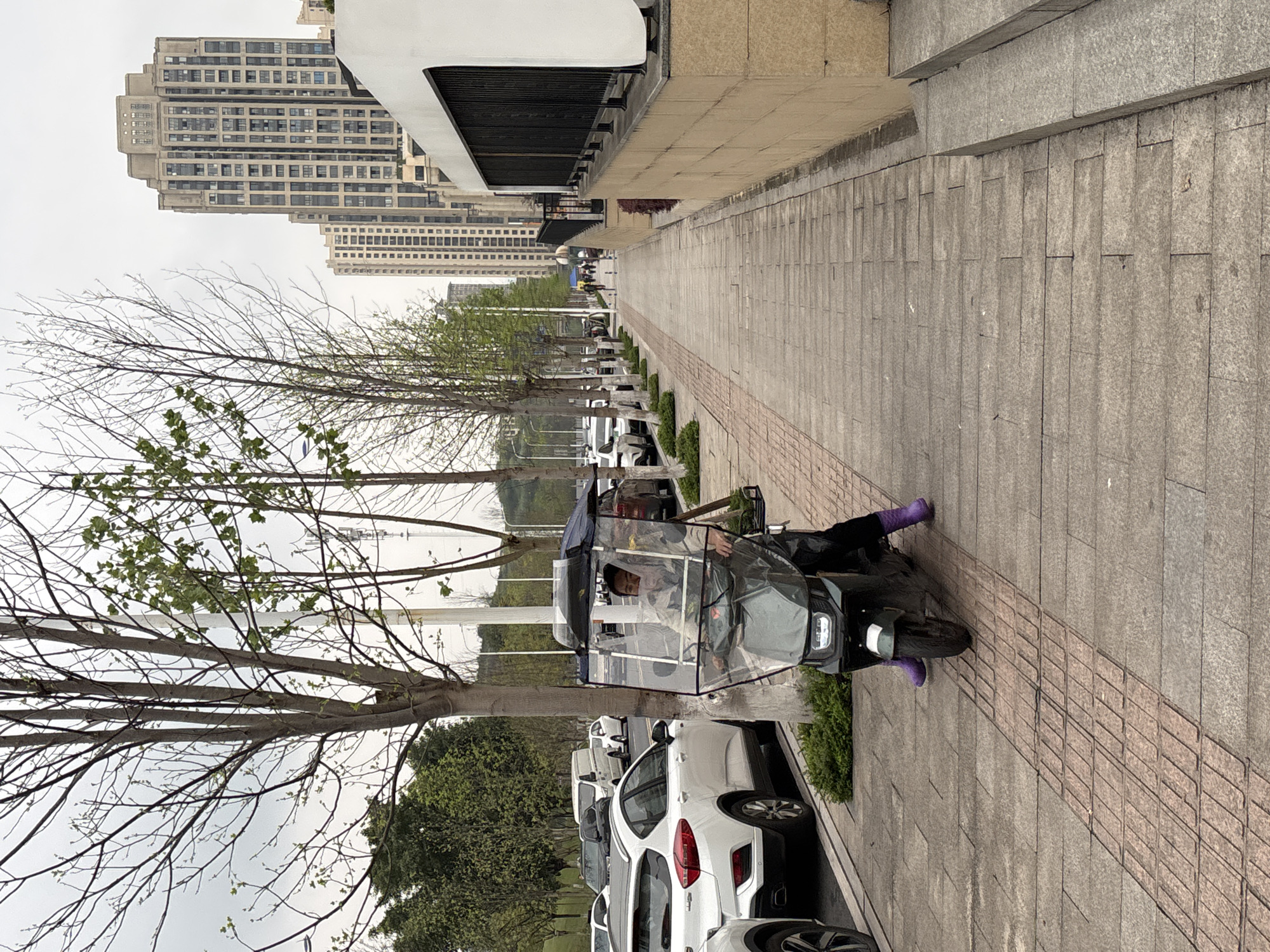 The scene depicts an urban street in Jiangjin, China, under an overcast sky. An individual, wearing a light-colored jacket and distinctive purple boots, operates a dark green electric scooter. The scooter is outfitted with a clear plastic canopy/windshield and a roof structure, and features a visible front headlight and a rear side-mounted carrier. The scooter is positioned on a paved sidewalk composed of grey and reddish-brown rectangular tiles, including a central strip of tactile paving. To the left of the sidewalk, a row of parked vehicles, primarily white, lines the street, with a white SUV prominent in the foreground. A series of slender trees with budding branches and sparse green leaves runs parallel to the sidewalk, separating it from the street. On the right, a tan-colored retaining wall with black metal railings extends along the sidewalk, featuring a set of stairs. In the background, multiple tall, modern apartment or office buildings rise above the surrounding structures. A few distant figures are visible walking further down the sidewalk.