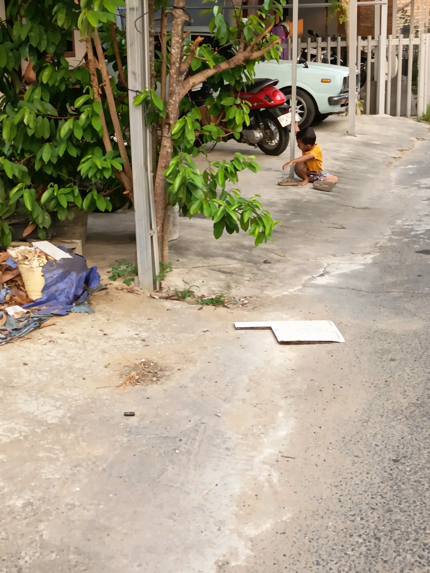 In Banjarmasin, Indonesia, a young boy in an orange shirt and patterned shorts squats on a concrete surface. To his right, a light blue car is parked behind a red scooter. A white fence runs along the background, partially obscured by greenery. A pile of debris, including a blue tarp, lies to the left of the boy, near the base of a tree. A piece of white cardboard is placed flat on the ground in the foreground.