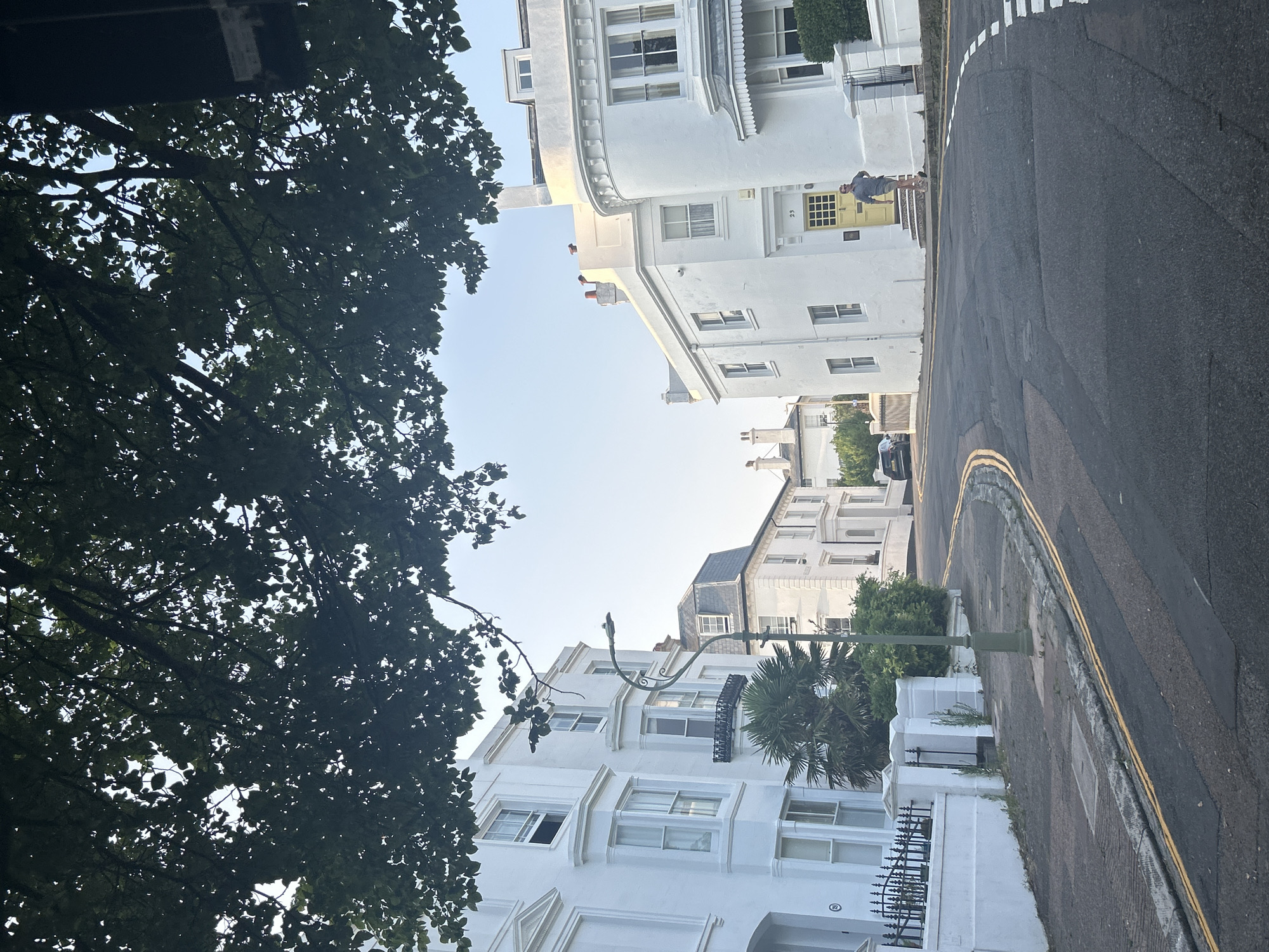 The image shows a street scene in the city of Brighton, United Kingdom, under a canopy of green leaves. The street curves to the right, with white buildings lining both sides. On the left, a white building with bay windows and black railings is visible. Further down the street, another white building with a gray mansard roof can be seen. On the right side of the street, a larger white building with a curved facade and a yellow door with the number "23" is prominent. A man in a blue shirt and tan shorts is walking out of the yellow door, holding a walking stick. The sky is clear and bright. The road has double yellow lines and white dashed lines.