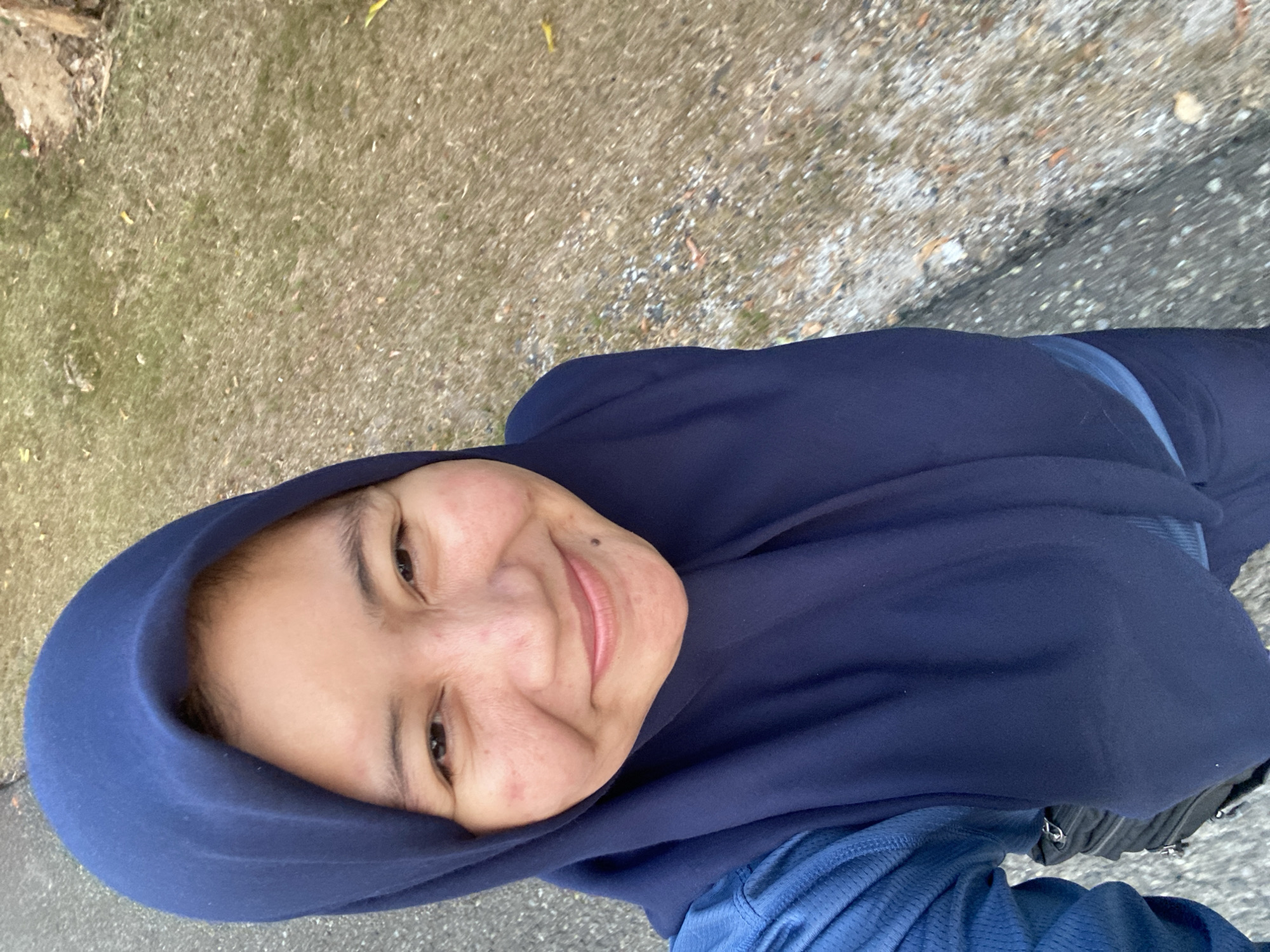 In Banjarbaru, Indonesia, a woman wearing a dark blue hijab and a blue shirt poses for a selfie outdoors. She has a slight smile and her face is turned towards the camera. The background is a mixture of asphalt and dry grass, suggesting she is near a road or pathway. A black bag is visible on her left side. The lighting indicates it is daytime.