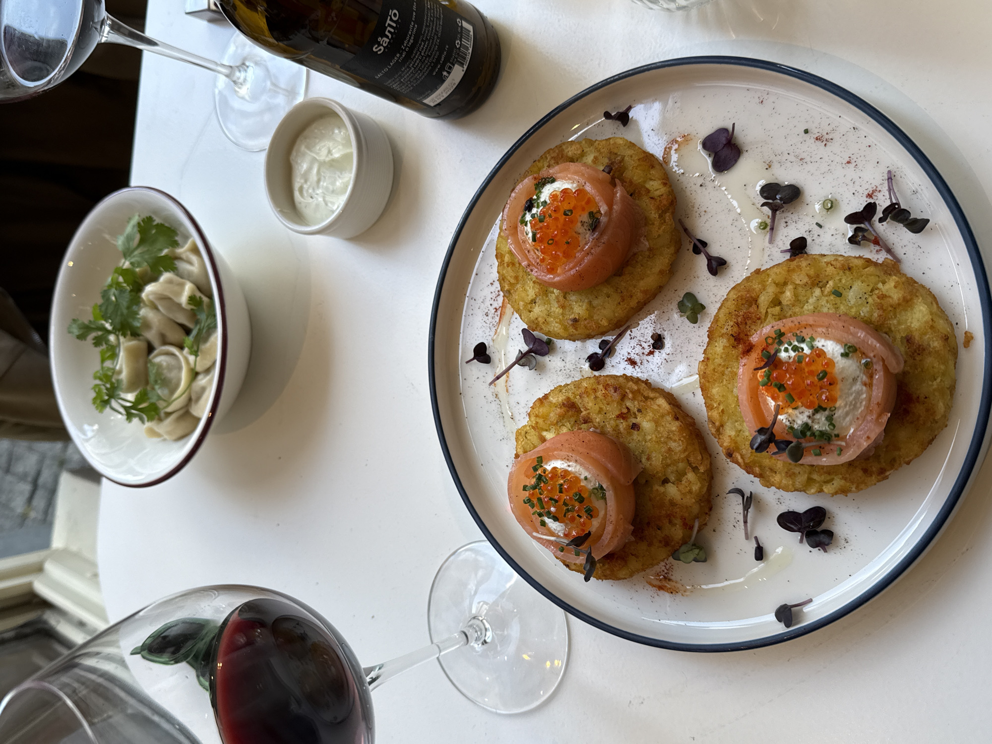 This high-angle shot captures a spread of dishes and drinks on a white table, indicative of a dining experience. The central focus is a white plate with a dark blue rim, presenting three round, golden-brown potato pancakes (likely latkes). Each pancake is artfully topped with a dollop of cream cheese or sour cream, a delicate curl of smoked salmon, and a spoonful of vibrant orange caviar, garnished with fresh chives. The plate is further adorned with scattered purple microgreens and red spice.
To the left, a small white bowl with a brown rim holds several light-colored dumplings, possibly pelmeni, topped with fresh green herbs like parsley. Beside it, a small white cup contains a creamy white sauce, likely intended as an accompaniment for either the pancakes or the dumplings.
Beverages visible include a dark glass bottle with a "Såntó SANTOLAGER" label, suggesting a craft beer, and two wine glasses. One glass, prominently in the bottom left, contains red wine, while another empty glass is partially visible at the top left. The bottle label text, partially legible, includes phrases like "Laboravite sve što ste znali o lagerima," "mutno, nefiltrirano, nepasterizovano," and "www.salto.rs."
The bright lighting and clean presentation suggest an indoor setting, such as a restaurant or cafe in Belgrade, Serbia, during the daytime. The arrangement implies a relaxed meal for one or two people.