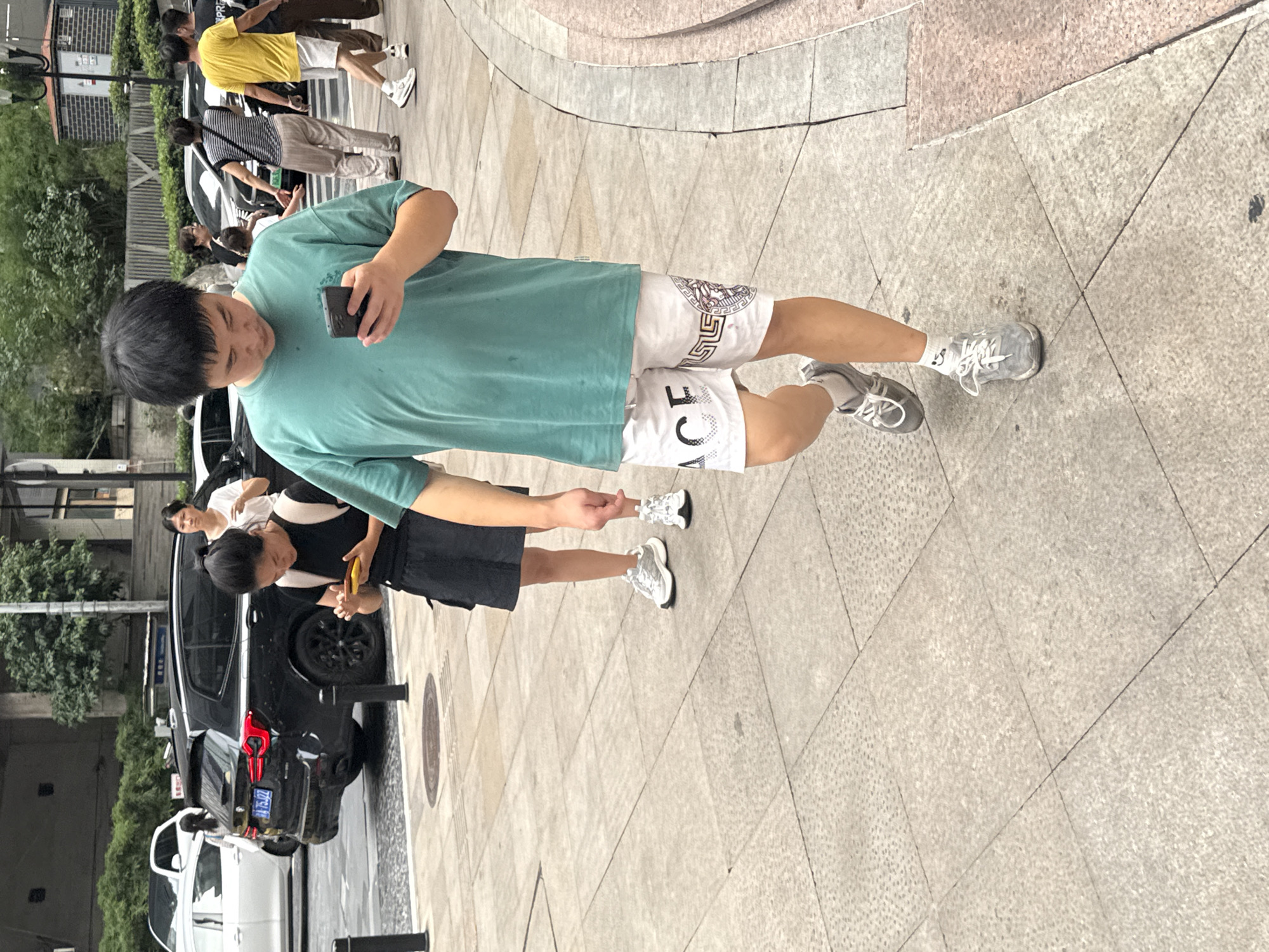 In Yuzhong District, China, on a pedestrian sidewalk paved with light-colored square tiles, a young man is walking while looking at his smartphone. He is wearing a teal t-shirt and white shorts with a Greek key pattern and a Medusa head logo. He has on gray athletic shoes and white socks. To his left, a young woman is also walking and looking at her smartphone. She is wearing a black sleeveless top, a black skirt, and white athletic shoes. In the background, several vehicles are parked on the street, including a black SUV. Trees and buildings are visible behind the vehicles, and a street lamp is present on the right side of the image.