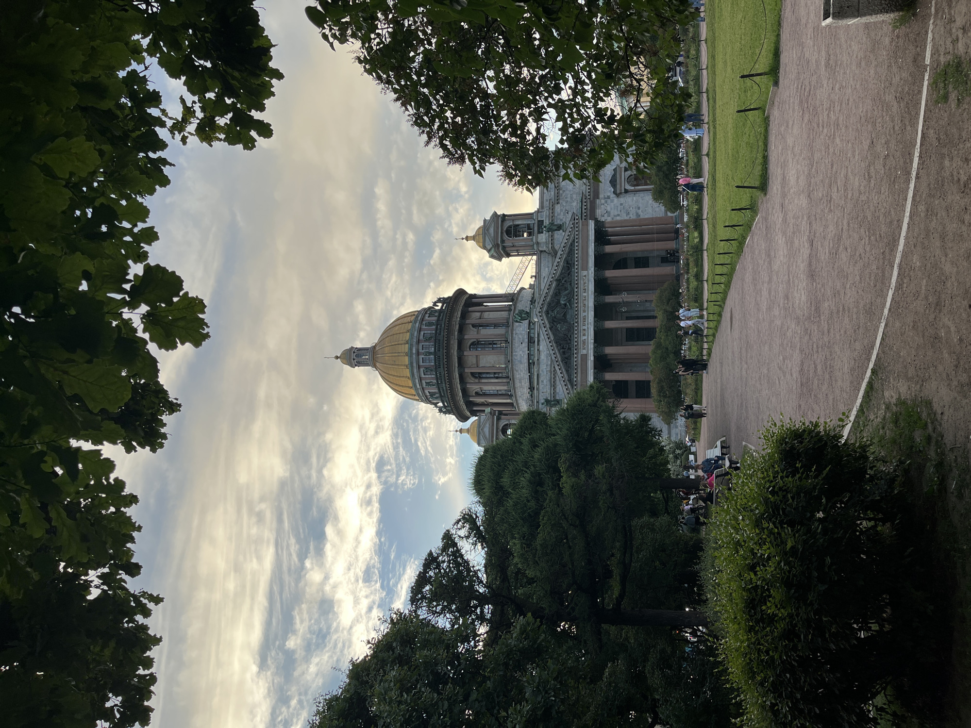This is a vertical, low-angle, outdoor photograph of St. Isaac's Cathedral in Saint Petersburg, Russia. The cathedral, with its distinctive golden dome, is partially obscured by the leafy branches of trees in the foreground. The sky above is partly cloudy, with streaks of white and grey clouds against a pale blue backdrop, suggesting either early morning or late afternoon light.
To the right of the cathedral, a gravel path winds through a green lawn bordered by a low fence. Several people are visible walking or sitting in small groups on benches along the path and on the grass. The atmosphere appears relaxed and leisurely, with people enjoying the outdoor space. The architecture of the cathedral itself, with its grand columns and ornate detailing, is impressive, even when partially framed by the natural elements. The sunlight filtering through the clouds casts a soft glow on the scene. No visible text is present in the image.