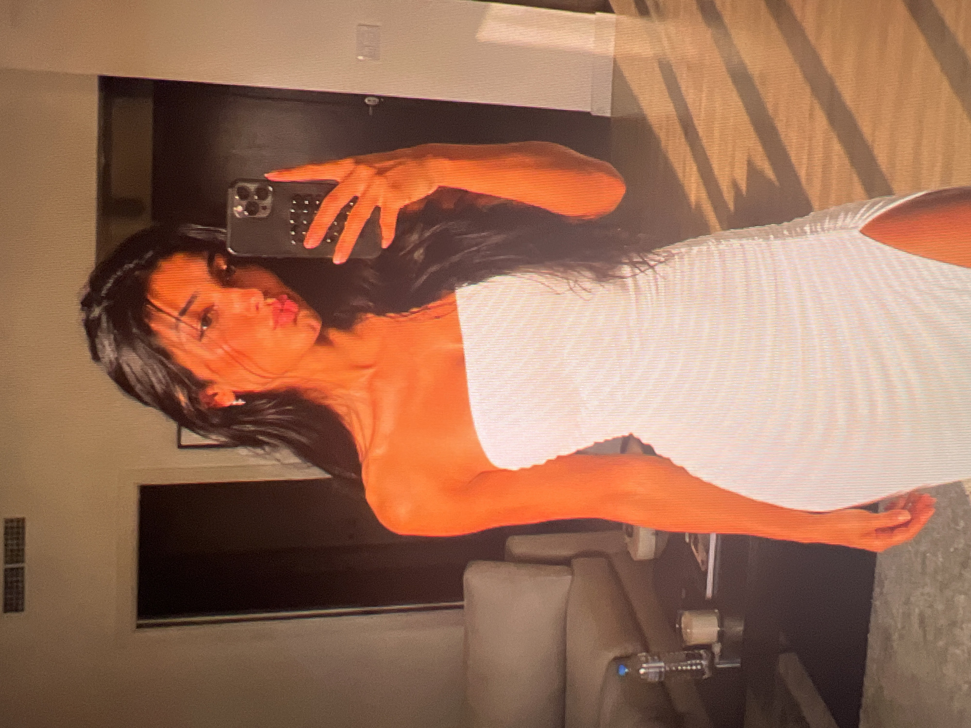 In the city of Unknown, Unknown, a young woman with long, dark hair takes a mirror selfie in what appears to be a modern apartment. She is wearing a strapless white ruched dress with a thigh-high slit. The lighting suggests it is late afternoon, with strong shadows cast across the wooden floor. In the background, a couch and a side table with a water bottle and a candle are visible. The woman is pouting for the camera, holding up her phone to capture the reflection.