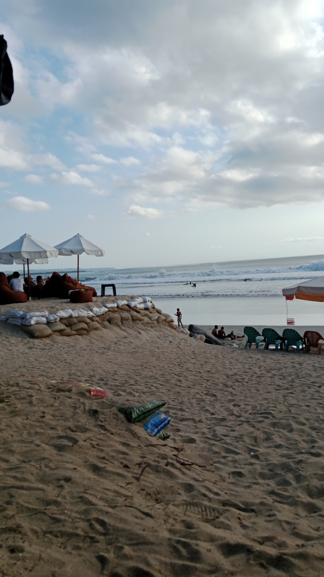 This is a medium shot of a beach scene in Kuta, Indonesia, likely during the late afternoon or early evening, given the lighting and the presence of surfers. The foreground is dominated by a sandy beach with some debris including plastic bags. In the midground, a sandbag barrier is visible, separating the beach from a seating area. This seating area features several large, beanbag-like chairs and two white umbrellas, with some people seated. Beyond the barrier and seating area is the ocean with moderate waves. Several surfers are visible in the water, engaged in the activity of surfing. The background shows a cloudy sky with patches of blue. To the right of the midground, a line of plastic chairs is set up, suggesting a casual dining or lounging area. The overall atmosphere is one of a relaxed beach environment with recreational activities taking place.