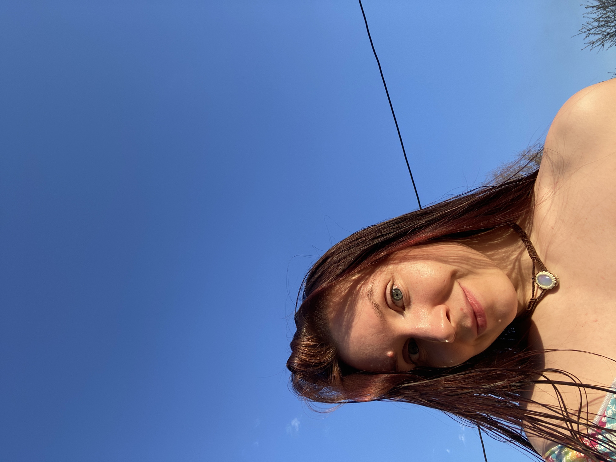 The image shows a woman with auburn hair and a necklace against a clear blue sky. A black cable runs diagonally across the top right of the frame. In the extreme upper right is a small cluster of tree branches. The woman is the primary subject of the image, with a focus on her face and upper body. The background is a cloudless sky, suggesting a sunny day. The location within the city of Фоки, Russia, cannot be determined from the visual information provided.