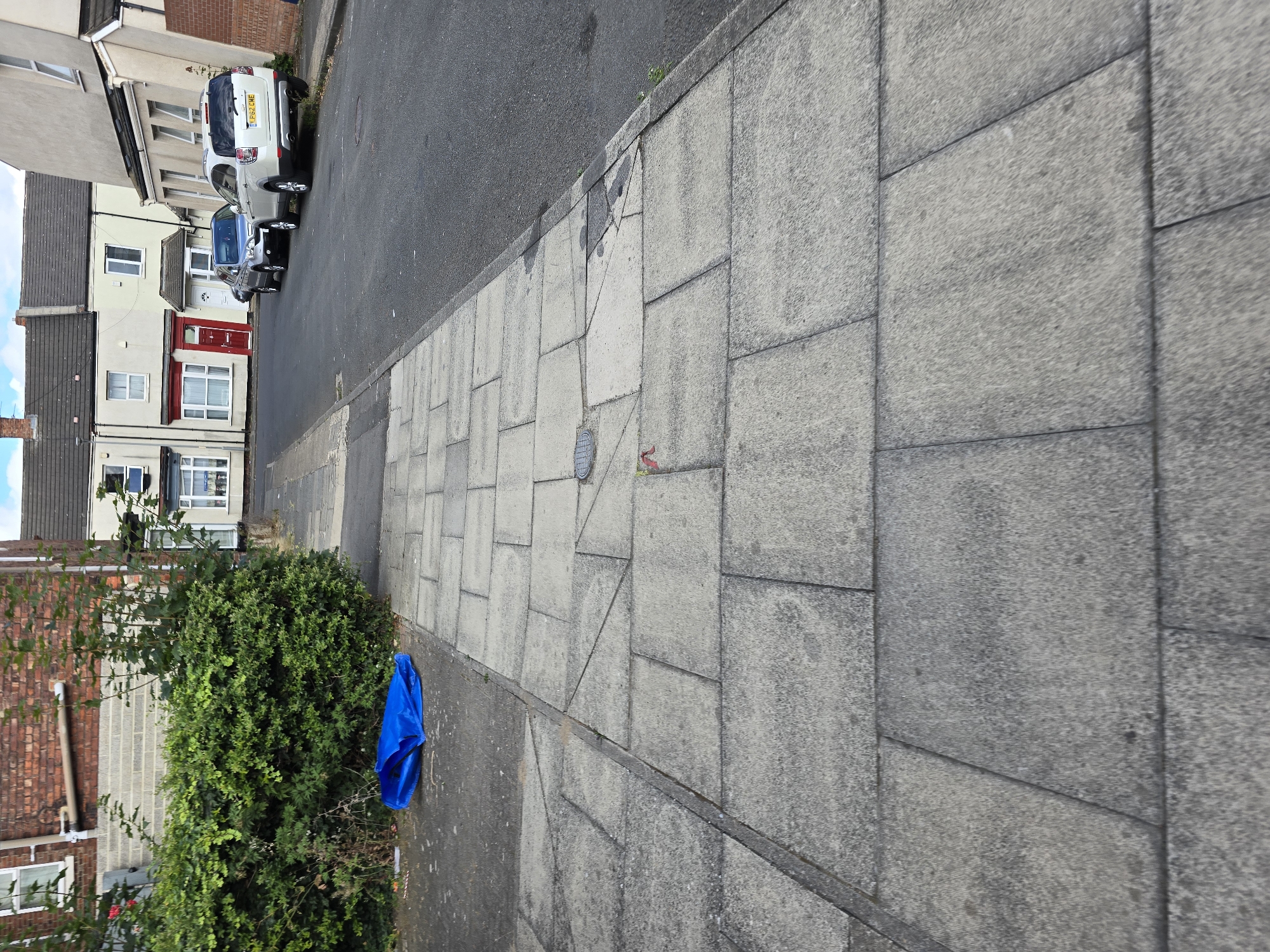 The image depicts a residential street in Stockton-on-Tees, United Kingdom, characterized by terraced houses and a paved sidewalk. In the foreground, the concrete paving slabs of the sidewalk are visible, showing some wear and tear. To the left of the sidewalk, a dense green hedge is situated against a brick building. A bright blue plastic bag lies on the ground near the hedge. The street itself is asphalt and several cars are parked along the curb. The houses in the background are typical of the area, with white and cream facades and dark tiled roofs. The sky is partly cloudy.