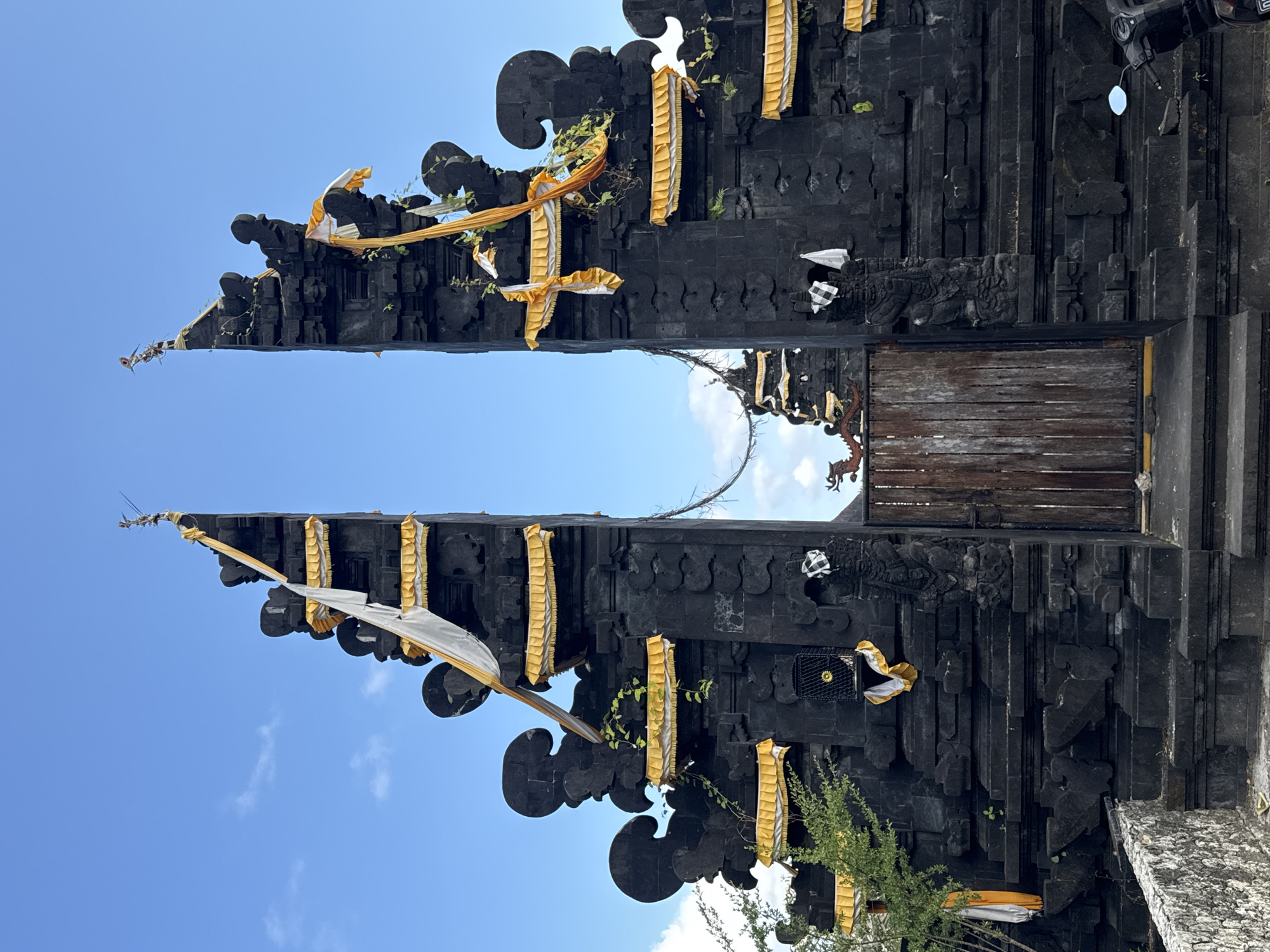 The image captures a traditional Balinese Candi Bentar (split gate) under a bright, clear blue sky in Kuta Selatan, Indonesia. The monumental gate, constructed from dark, possibly volcanic stone, features intricate carvings and stands as a prominent architectural structure.
Numerous decorative elements adorn the gate, including flowing yellow and white/silver fabrics and banners draped over its multi-tiered spires and along its horizontal sections. Green vines and small plants are seen growing on the dark stone, suggesting age and integration with nature. Between the two towering halves of the gate, a rustic wooden door or panel is visible on the right side. On the right section of the gate, a small, distinctive black and white checkered fabric, known as Poleng, is tied, accompanied by a small, dark, cage-like object with a yellow-gold circular center. A glimpse of a motorbike's handle and mirror is just visible at the top right edge of the frame.
The scene is static, highlighting the architectural detail and cultural significance of the gate without any human activity. The clear blue sky and strong illumination indicate a sunny day. No discernible text is visible in the image.