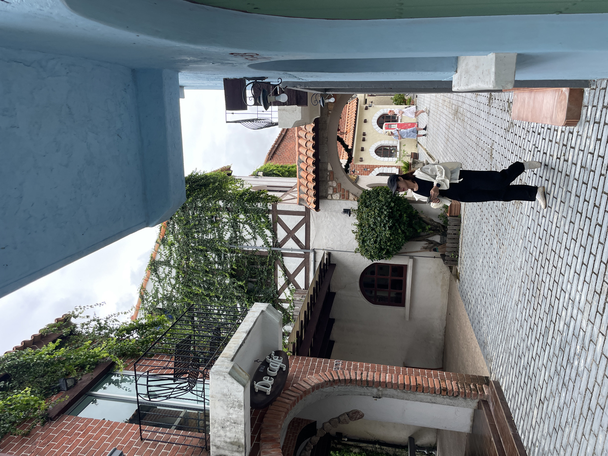 A woman wearing a black cap, black shirt, white jacket, black pants, and white sneakers walks through a cobblestone alleyway, holding a phone. She is in the foreground, walking from left to right. The alleyway is lined with buildings featuring brick and white facades, some adorned with ivy. A sign with "De café" is visible on one of the buildings. In the background, two children, a boy and a girl, are standing in the alley. The scene appears to be an outdoor setting in a European-style village, possibly part of a themed attraction in Na Chom Thian, Thailand. The sky is overcast.