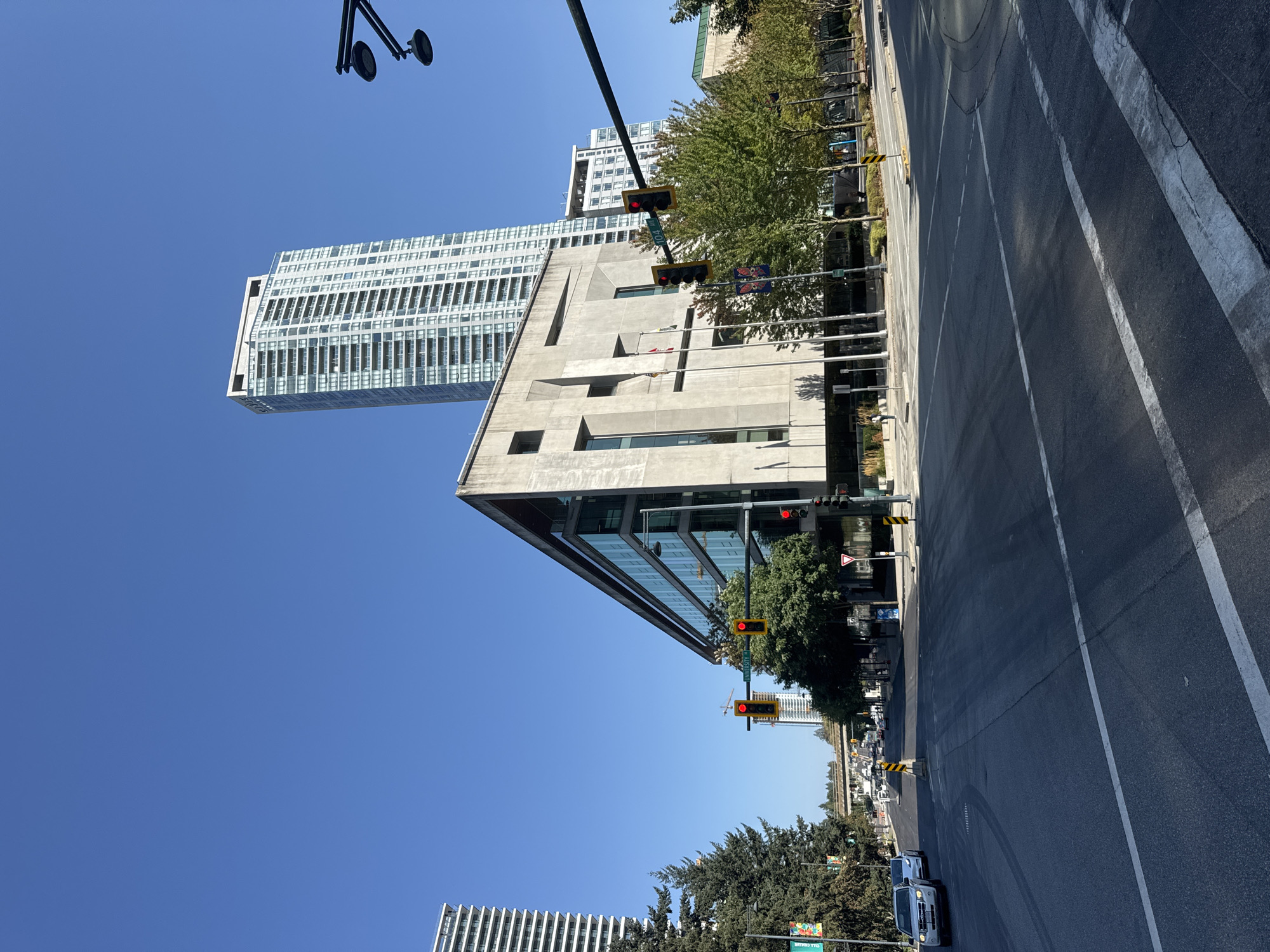 This is a daytime, eye-level, medium shot of a street intersection in Surrey, Canada, specifically at the intersection of University Drive and 104 Avenue. The scene is dominated by a large, modern concrete building with glass windows on the left side of the frame. In the background, a tall, slender, light-colored residential skyscraper with numerous balconies rises into a clear blue sky.
To the right of the concrete building, several trees with green foliage line the sidewalk. Traffic lights are visible at the intersection, with red lights illuminated. Cars are present on the road, with one white SUV visible in the foreground left. Street signs, including one indicating "104 AVE", are also present. The road surface is asphalt with white lane markings. The overall environment appears to be an urban setting with a mix of modern architecture and greenery. There are no individuals clearly visible in the image.