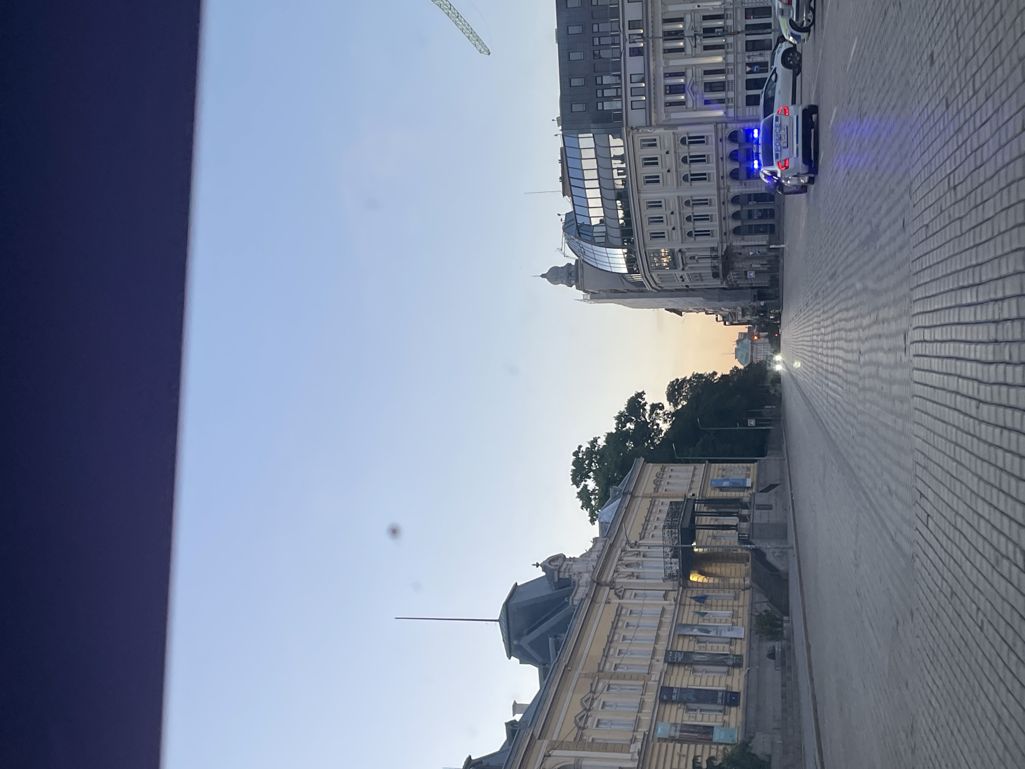 The image captures a street view in Sofia, Bulgaria. A street is shown with brick pavers. On the right side of the street, police vehicles with flashing blue lights are parked. Buildings line both sides of the street, including a taller, modern building with glass and a traditional, yellow building. Trees are visible. The sky is light blue with hints of orange, suggesting either sunrise or sunset. A dark, solid object is on the left edge of the frame, obscuring part of the sky.