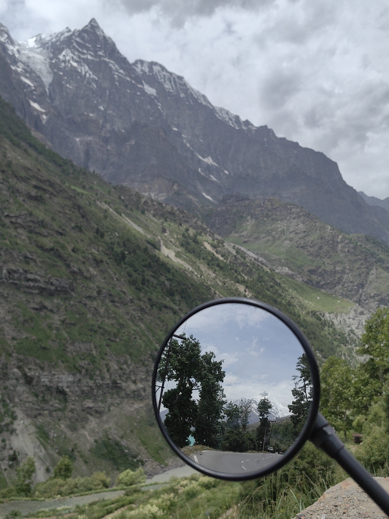 This image captures a scenic mountain landscape, likely in or near Jagle, India, as reflected in a motorcycle's side-view mirror. The foreground is dominated by the mirror, mounted on a black stem, which offers a distorted but clear reflection of the road and the surrounding scenery.
Within the mirror's reflection, a winding asphalt road is visible, bordered by lush green trees. In the distance, a snow-capped mountain peak stands majestically against a cloudy sky. The mirror also shows a few scattered trees and possibly distant buildings or structures.
Outside the mirror, the actual landscape is revealed. A steep, green mountainside fills the left side of the frame, with evidence of terraced fields or agricultural plots at its base, near a flowing body of water. The upper portion of the frame is occupied by a towering, rugged mountain range, its slopes covered in sparse vegetation and patches of snow. The sky above is overcast with heavy clouds, suggesting a cool or potentially rainy day. The overall impression is one of a remote, natural environment with dramatic geographical features.