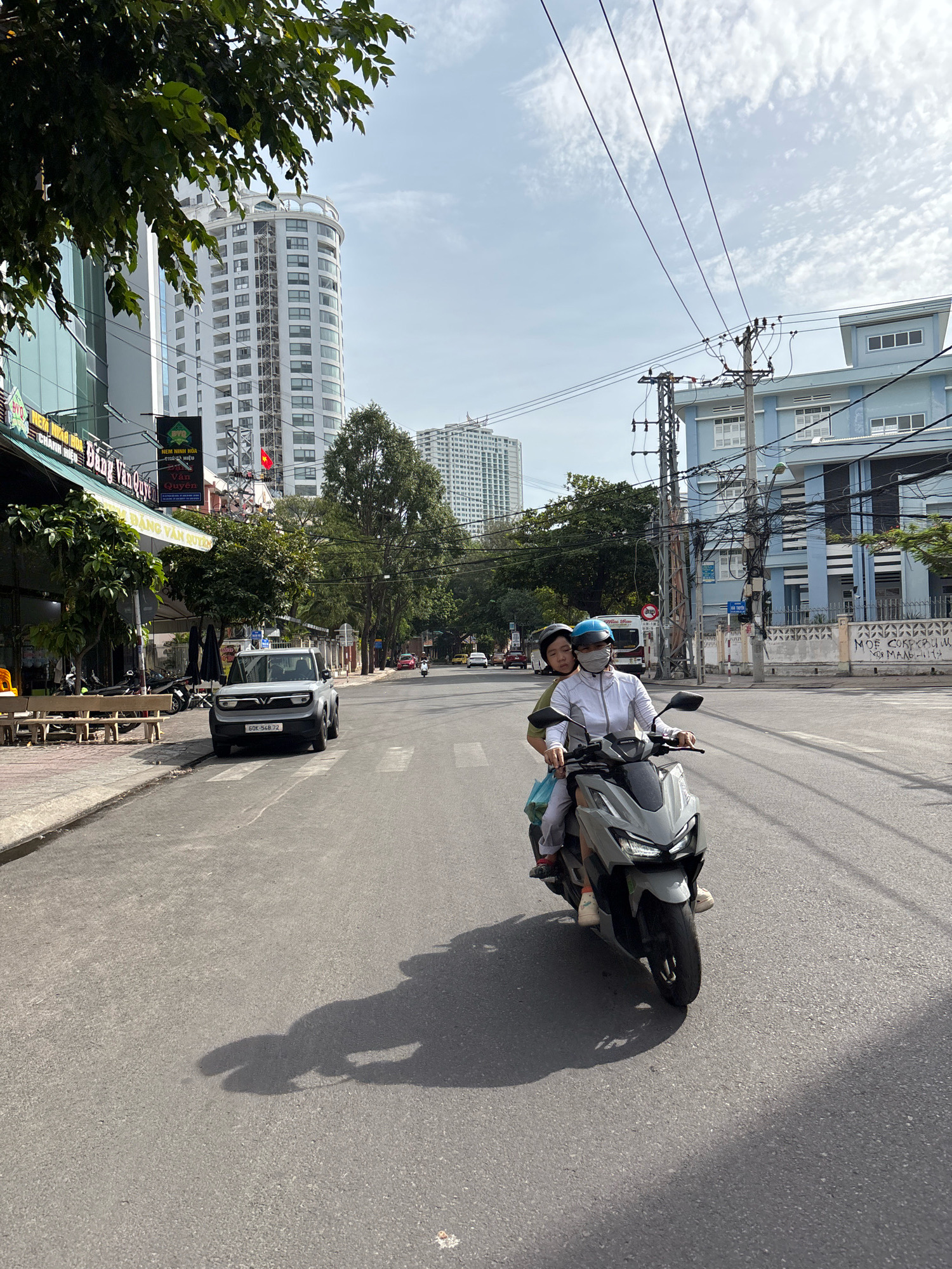 Two individuals, likely adults, are riding a grey scooter on a paved street in Nha Trang, Vietnam. The person in front is driving, wearing a white long-sleeved shirt, a face mask, and a blue helmet. The person behind is wearing a green shirt and a black helmet. They are both wearing casual clothing. The scooter is moving forward on the right side of the road, casting a shadow on the asphalt. In the background, there is a light blue building with multiple windows and a complex network of electrical wires and poles. To the left, a white VinFast car is parked on the side of the road, near a sidewalk with benches and trees. Further in the distance, tall white buildings are visible, characteristic of urban development in Nha Trang. The sky is partly cloudy. The scene appears to be a typical urban street in Vietnam during daylight hours.