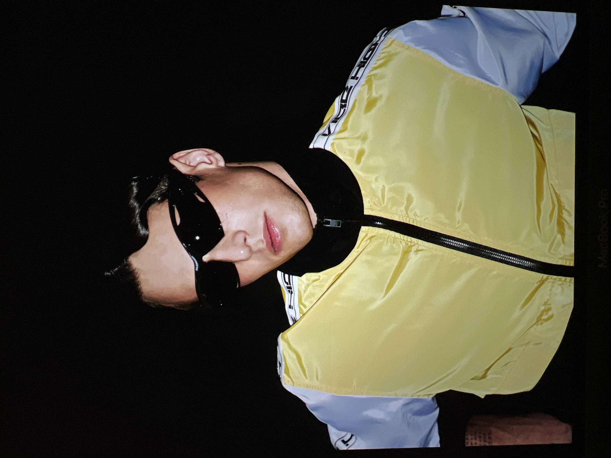 A medium shot captures a young man wearing sunglasses and a yellow and white jacket with black trim against a black background. The man's hair is styled with a pompadour. He is looking directly at the camera. The jacket has a zipper down the front, a black collar, and white stripes with text on the shoulders. The background is entirely black, creating a studio portrait feel. There is no visible indication of location within a city or country.
