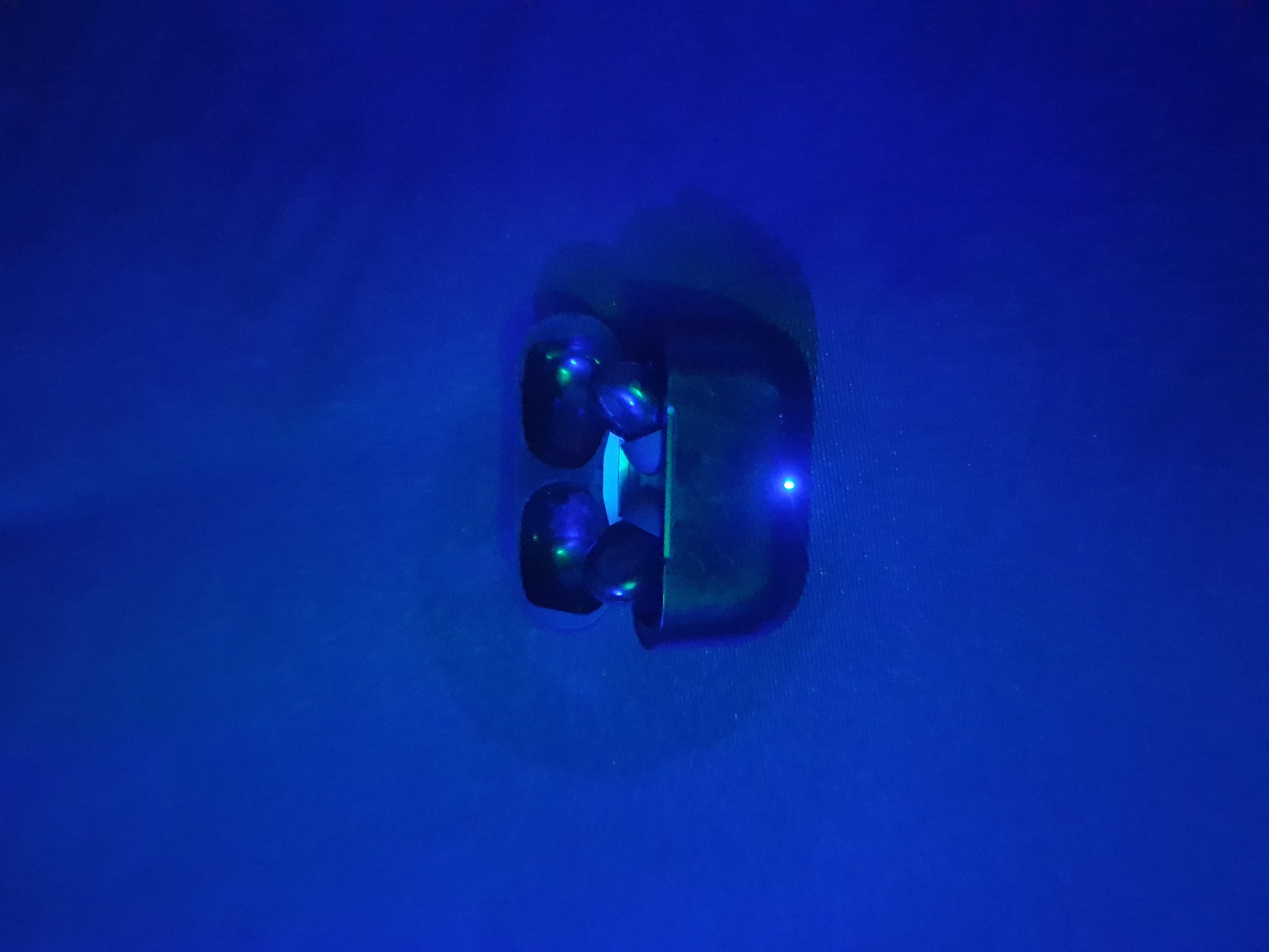 An overhead shot shows a pair of black wireless earbuds resting in their charging case. The earbuds are partially visible, with hints of green light reflecting off their surfaces. A small blue LED light is illuminated on the front of the charging case. The entire scene is bathed in a deep blue light, creating a dramatic and somewhat mysterious atmosphere. The background is a solid, dark blue surface with subtle variations in shade and texture. There are no people or other objects visible in the image. The earbuds and case appear to be positioned on a flat surface.