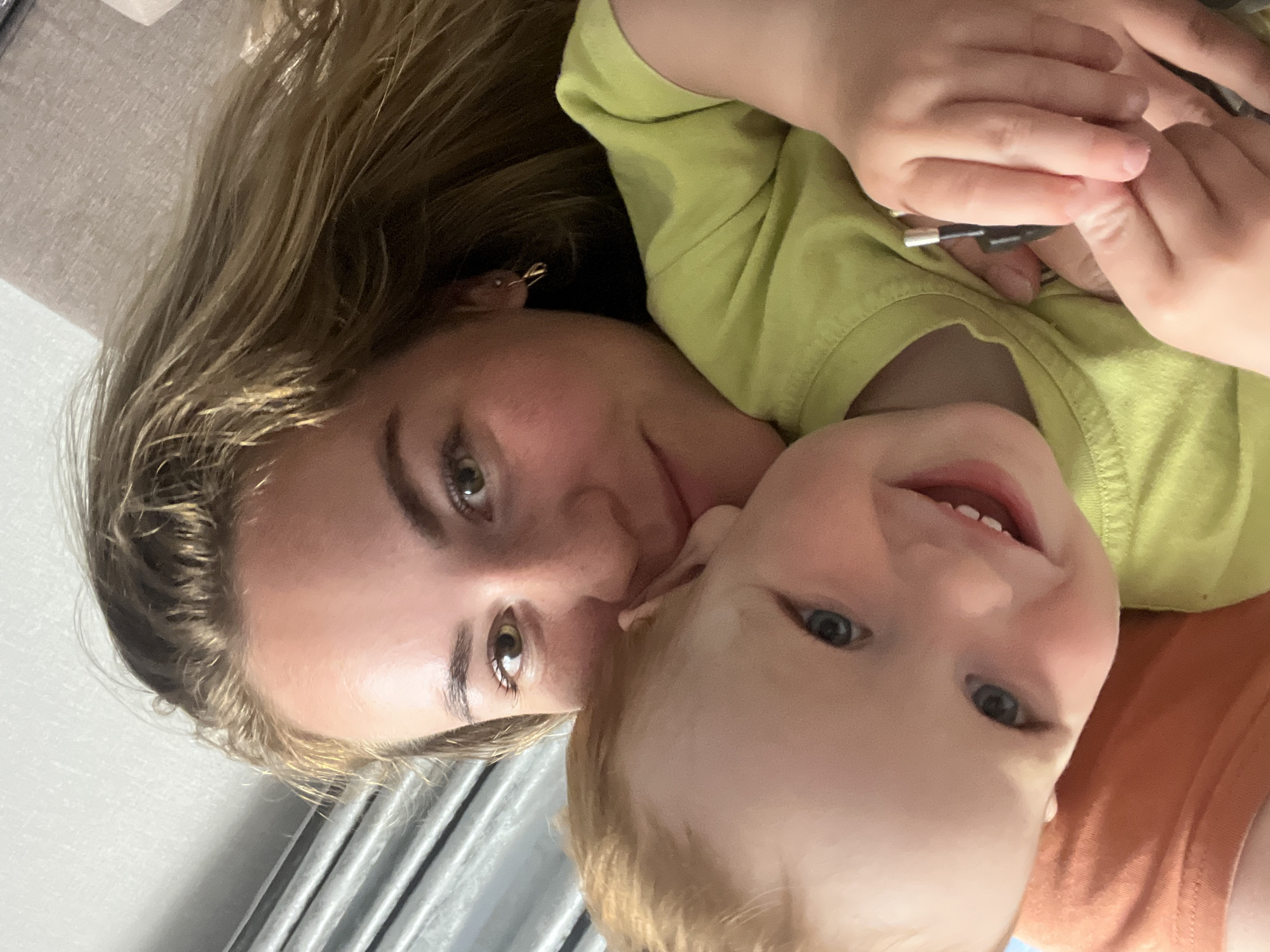 This is a close-up selfie of a young woman and a toddler. The woman has long, wavy blonde hair and is wearing a light green t-shirt and a gold earring. She is smiling and holding the toddler close to her. The toddler has blonde hair and blue eyes and is wearing a light green t-shirt. The toddler is smiling widely and appears to be looking at the camera. The background is blurry and appears to be indoors, possibly in a home. There are no discernable landmarks or contextual clues that would indicate the location in Chaikovsky, Russia.