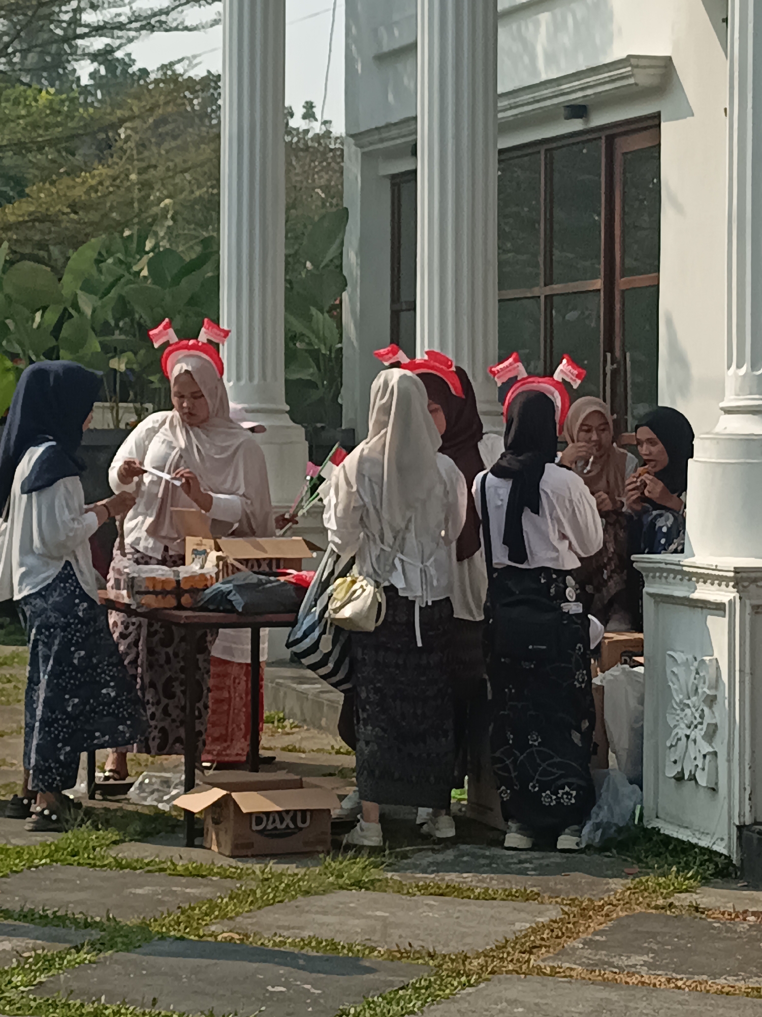 In Sleman, Indonesia, a group of women are gathered outdoors, with a white building and lush greenery in the background. The women, all wearing headscarves, appear to be engaged in social interaction and possibly a transaction. Several of them are wearing festive red and white headbands, suggesting a celebration or national event. One woman in the center is handing a white object, possibly a receipt or a small item, to another woman. A table in front of them is laden with what appear to be food items, perhaps snacks or sweets, displayed in trays and boxes. Another box, labeled "DAXU", sits on the ground near the table. The ground is paved with concrete and interspersed with patches of grass, and the sunlight indicates it is daytime.