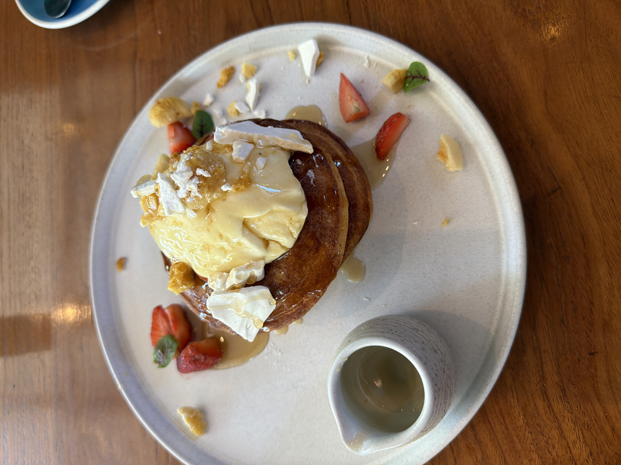 The image shows a plate of food, likely pancakes, topped with a yellow cream, meringue pieces, and strawberries. A small pitcher of syrup or sauce is next to the pancakes. The plate is on a wooden table, suggesting a restaurant setting. The location is unspecified, only that it is in the city of Unknown, Unknown.
