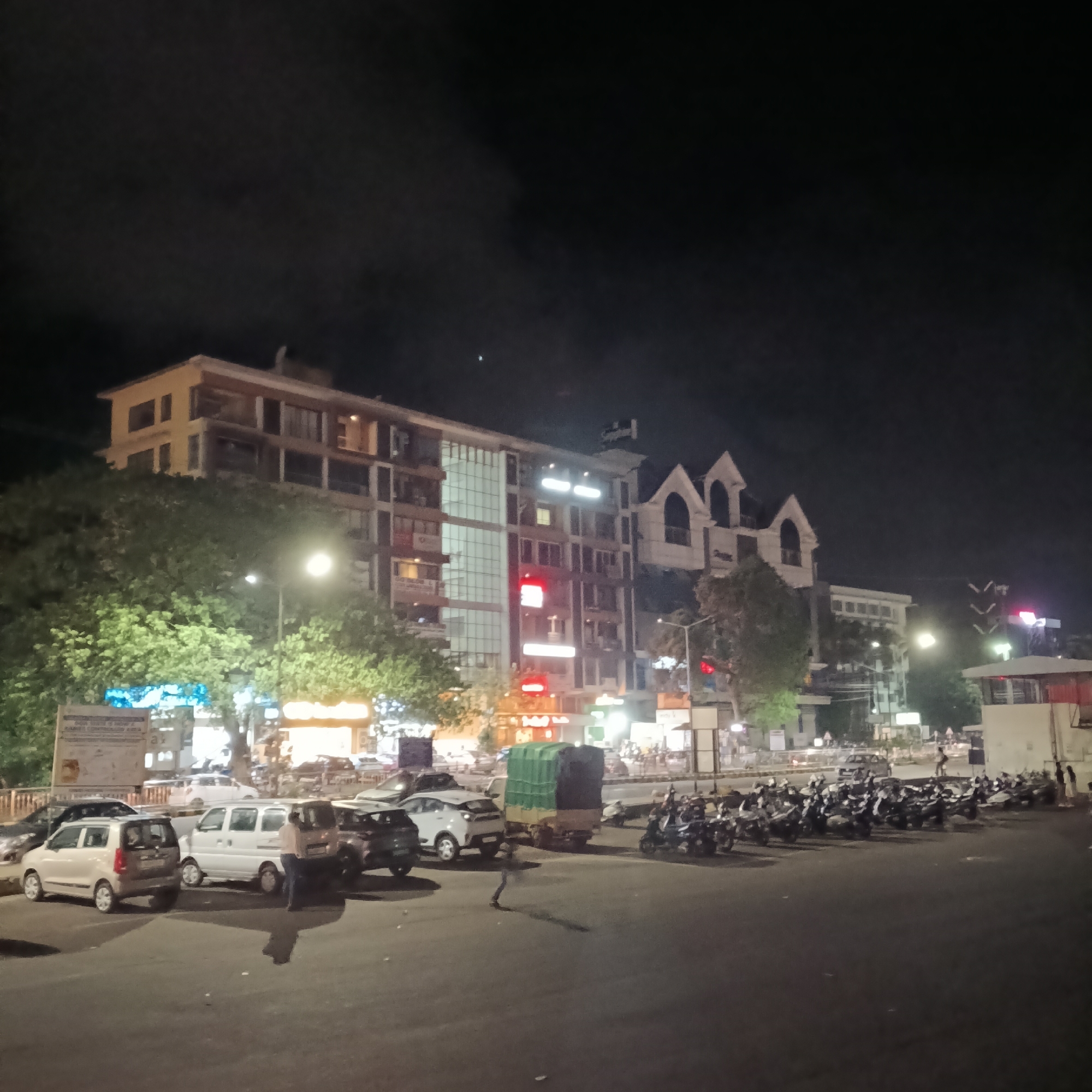 At night in Margao, India, a street scene shows cars parked on the left and rows of parked motorcycles on the right. Buildings are in the background, illuminated by streetlights and business signs. There are pedestrians. The road is cracked. The sky is dark.