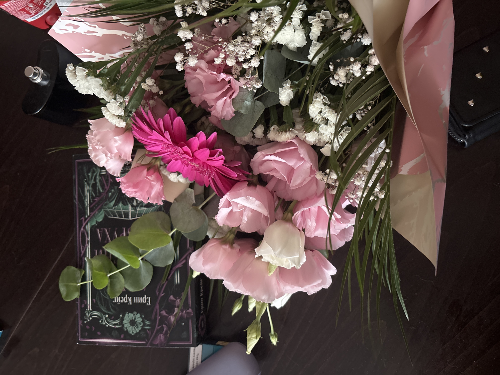 The image shows a close-up, overhead view of a bouquet of pink and white flowers, along with eucalyptus leaves, arranged on a dark wooden surface. The bouquet is presented with decorative pink and white paper wrapping. Partially visible behind the flowers is a black book with an ornate cover, featuring green and purple imagery and text in Cyrillic script. To the right of the book, a black flask-shaped object with a silver cap is visible, and further to the right, a red object, possibly a bottle or can, is also partially in frame. The overall scene suggests a gift or a decorative arrangement. There is no indication of the city of Veliko Tarnovo, Bulgaria, within the image.