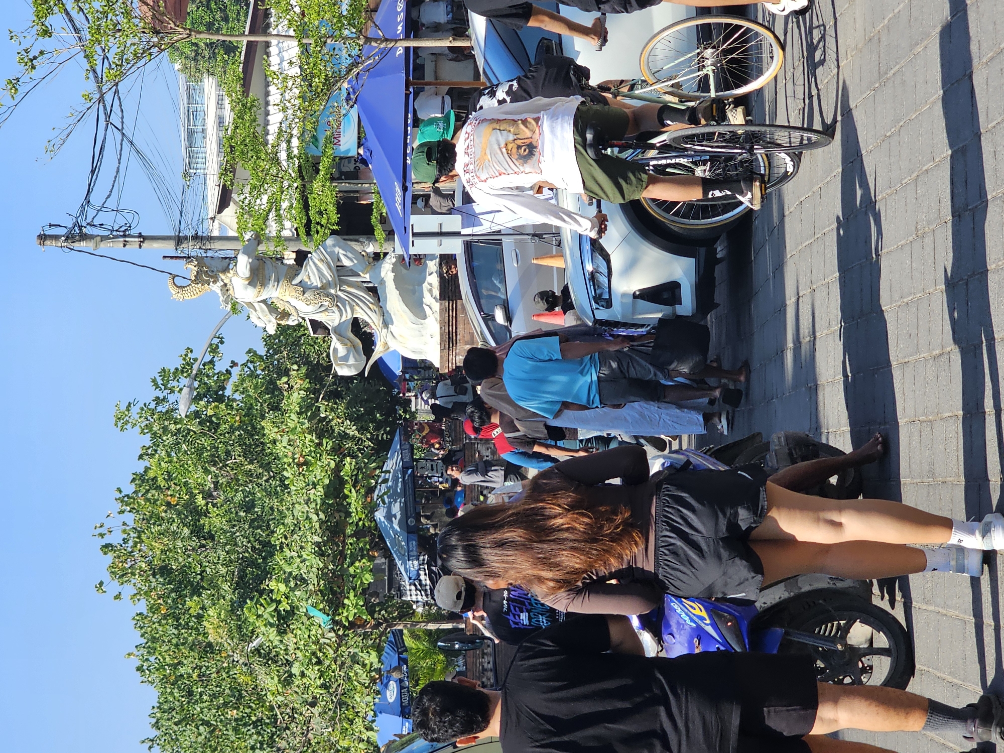 In Denpasar, Indonesia, a busy street scene unfolds under a bright, clear blue sky. A large, ornate white statue of a warrior figure stands prominently in the background, flanked by lush green trees. The street is paved with interlocking bricks and shows signs of vehicular traffic, with several cars parked or moving. Blue umbrellas, likely for vendors or cafes, are scattered throughout the area.
Several people are visible, including a woman in black shorts and a brown long-sleeved shirt in the foreground, standing near a blue and black motorcycle. A man in a black t-shirt is to her left, and another man in a white long-sleeved shirt with a graphic print is riding a bicycle towards the right. Other pedestrians are seen walking or standing, some carrying bags. The overall atmosphere appears to be that of a bustling urban environment, possibly a market or a tourist area, with a mix of locals and visitors.