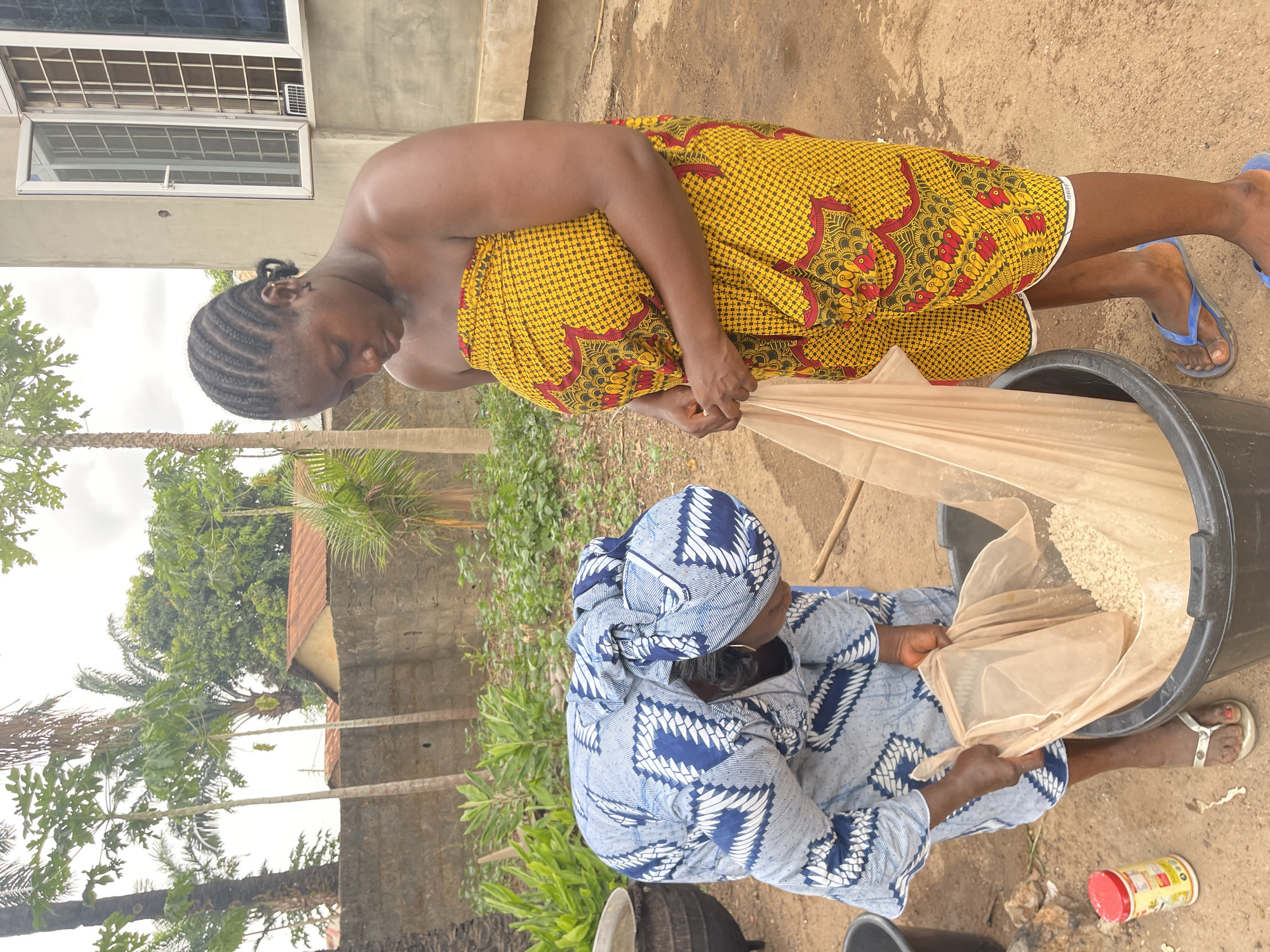 Two women are outdoors in Akwanga, Nigeria, processing grains. One woman, wearing a yellow patterned wrap, is standing and pouring grains from a white bag into a large black bucket. The second woman, wearing a blue and white patterned headwrap and outfit, is kneeling and holding the bag within the bucket. A small red and yellow container sits on the ground nearby. The surrounding environment includes trees, a wall, and a building.