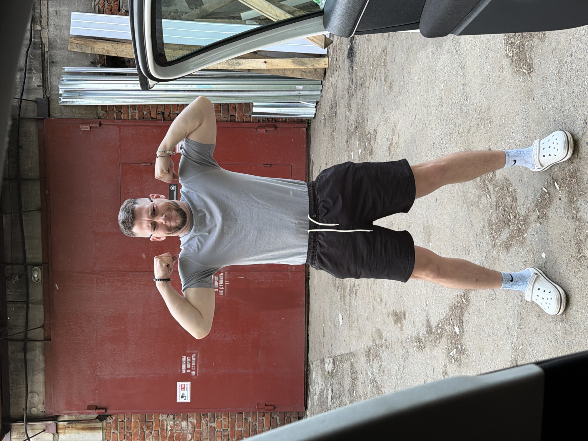 A man is posing outside a garage. He is wearing a gray t-shirt, black shorts, and light blue Crocs with socks. The background features a red brick garage door and what appears to be construction materials. The location is in an urban area with concrete ground and a vehicle is nearby.