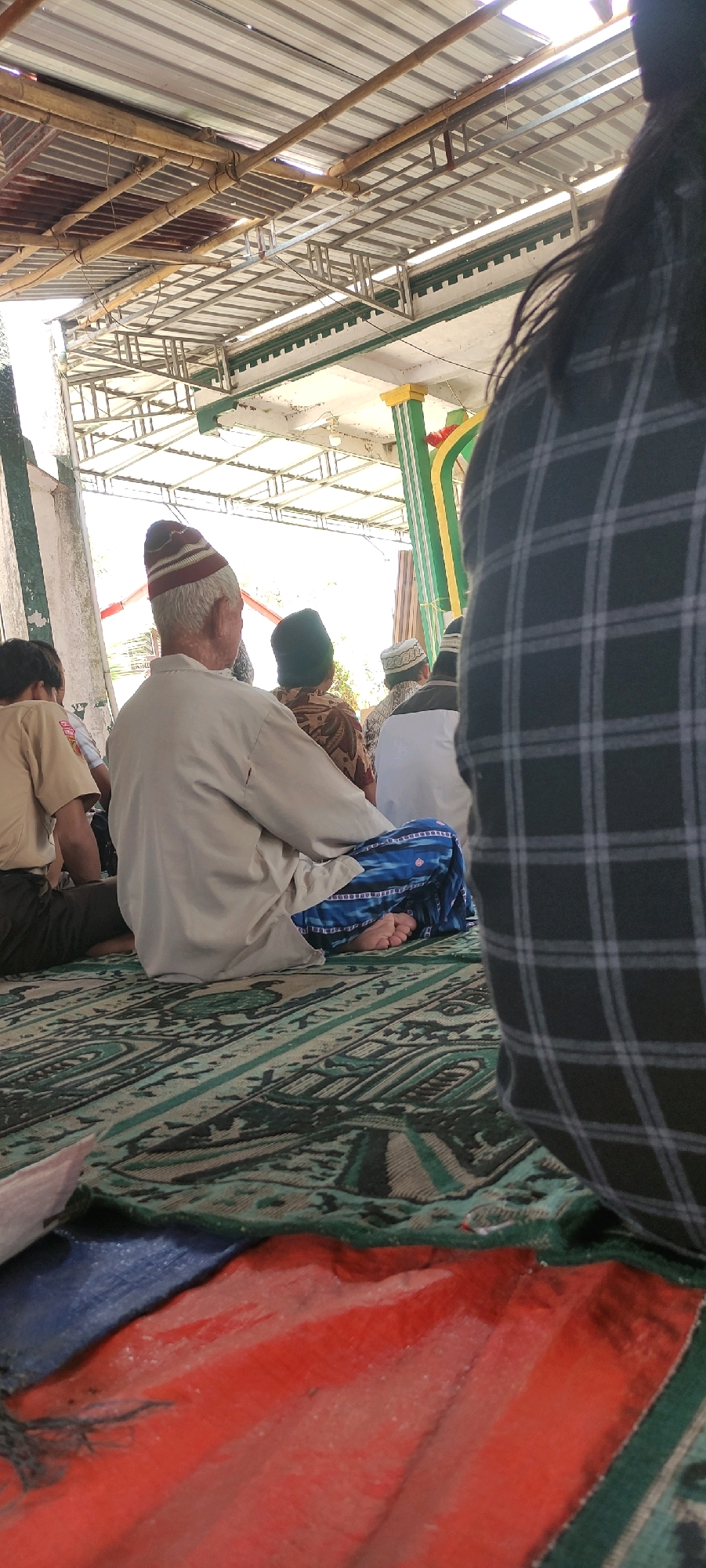 In Wadaslintang, Indonesia, the scene is set beneath a corrugated metal roof supported by bamboo and metal beams, suggesting an outdoor or semi-outdoor gathering. The focus is on a group of people seated on the floor, which is covered by a large, patterned carpet.
In the foreground, a person wearing a dark, plaid shirt is seen from behind, their shoulder and head obscuring a portion of the scene. To their left, an elderly man is seated, facing away from the camera. He wears a light-colored robe and a striped, conical hat. His legs are crossed, and he wears a blue and white patterned sarong, with one bare foot visible.
Behind the elderly man, other individuals are also seated. One person wears a dark green cap and a patterned brown and white shirt. Another, further back, wears a white and black cap and a white top. To the far left, a person in a tan shirt and dark pants is also seated. The overall impression is one of a religious gathering, likely prayer, given the seating arrangement and attire.
The environment is bright, with natural light filtering through the roof. The background reveals some architectural elements, including green and yellow striped pillars and what appears to be part of a building. The presence of multiple individuals suggests a community event.