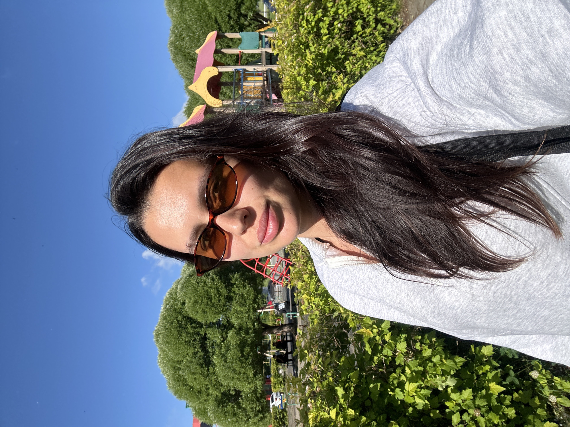 A woman with dark hair and sunglasses is taking a selfie outdoors. She is wearing a grey hoodie. Behind her, there is a playground with a slide and a climbing structure. Green bushes and trees are visible, along with a bright blue sky. The location is the city of Chaikovsky, Russia.