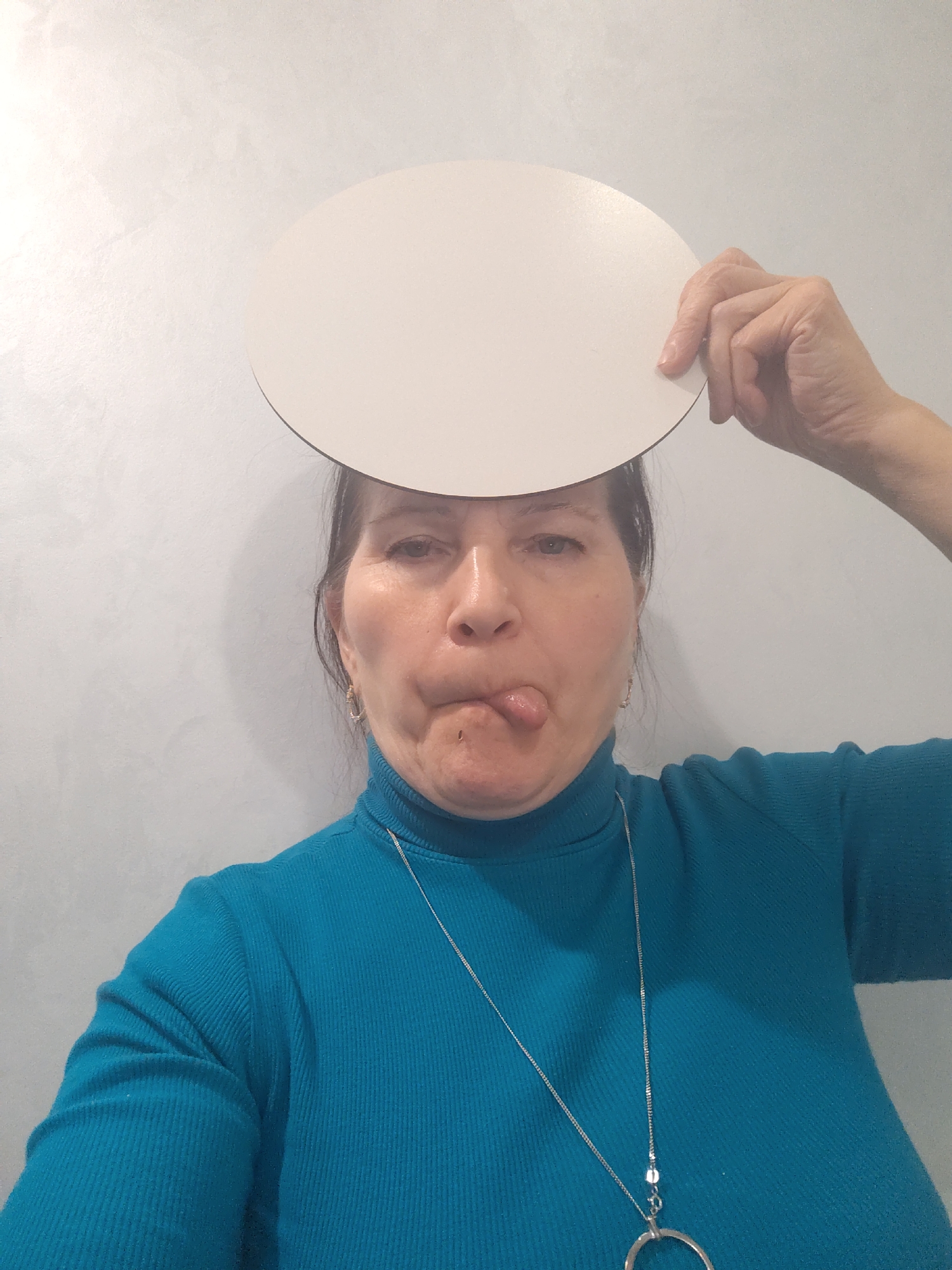 A middle-aged woman is depicted indoors, likely in Sarapul, Russia, making a playful expression. She is centered in the frame against a plain, light-colored wall that has a subtle, mottled texture. The woman has dark hair and is wearing a vibrant teal or turquoise ribbed turtleneck sweater. A long silver necklace with a large circular pendant and smaller metallic beads is visible draped over her sweater.
With her right hand, she holds a flat, plain white oval-shaped object above her head, partially obscuring her forehead. Her facial expression is humorous, with her tongue slightly sticking out to the side of her mouth. Her eyes are open and directed slightly upwards, contributing to the lighthearted mood. The lighting suggests an indoor setting, likely with artificial light, creating a soft shadow behind her head on the wall. No text is visible in the image.