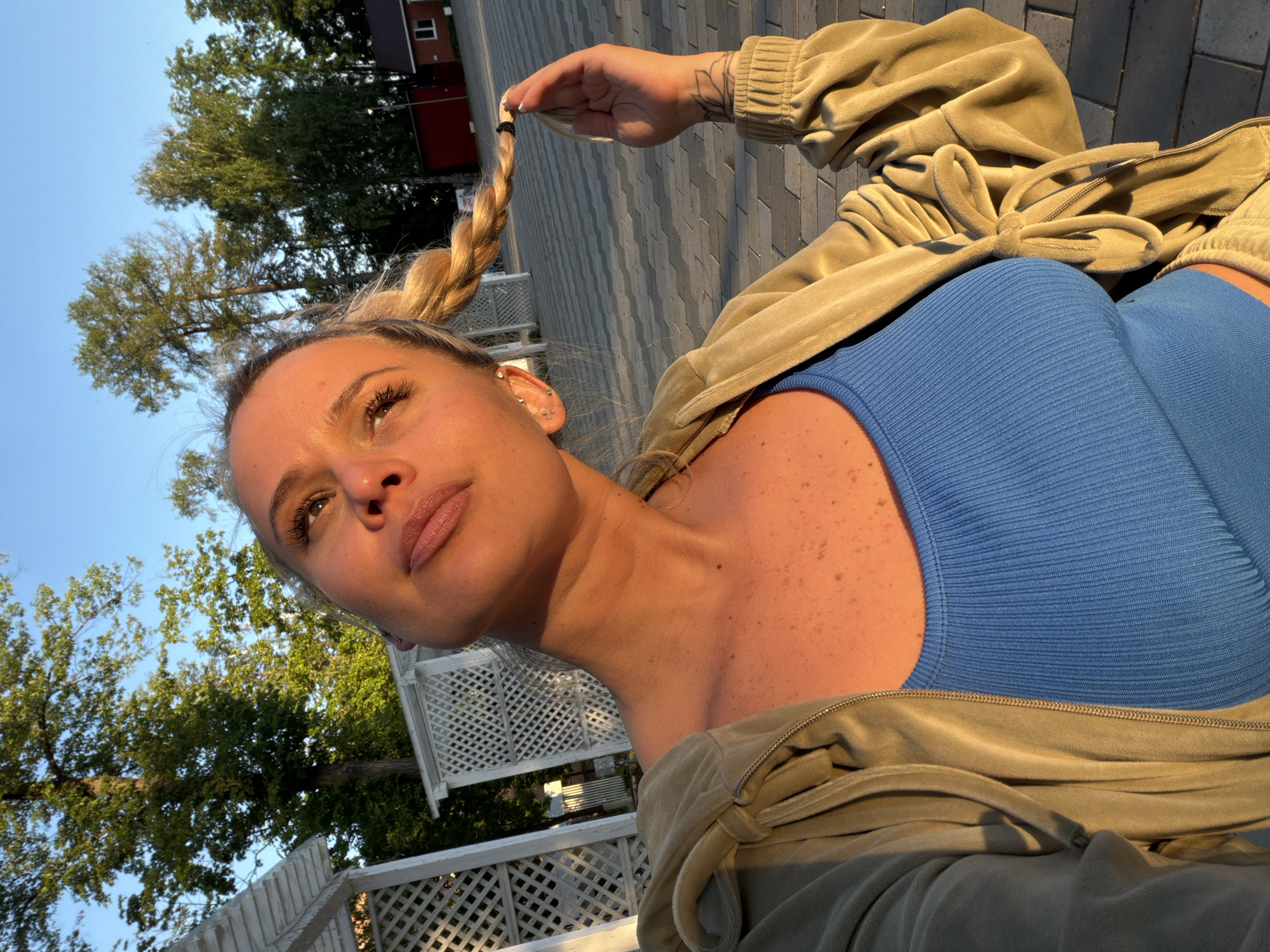 A woman is photographed from a low angle in what appears to be an outdoor urban setting. The woman has blonde hair, styled in a braid, and is wearing a blue top and a light brown jacket. She is holding the end of her braid with her left hand, which has a visible tattoo on the forearm. The background features trees, a building with a red roof, and a paved area with a geometric pattern. A white picket fence is also partially visible. The lighting indicates it might be late afternoon or early evening. Location: possibly in the city of Sarapul, Russia.