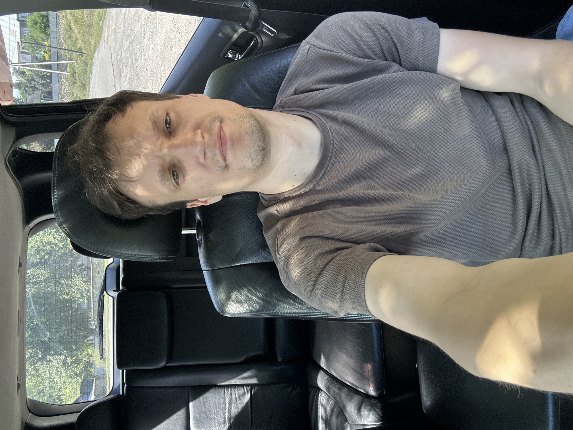 A man with brown hair and light green eyes sits in the driver's seat of a car, looking at the camera. He is wearing a brown t-shirt. The interior of the car appears to be black leather. The background shows a rural scene with trees, a fence, and a house. The image was taken in Makiv, Ukraine, on a sunny day.