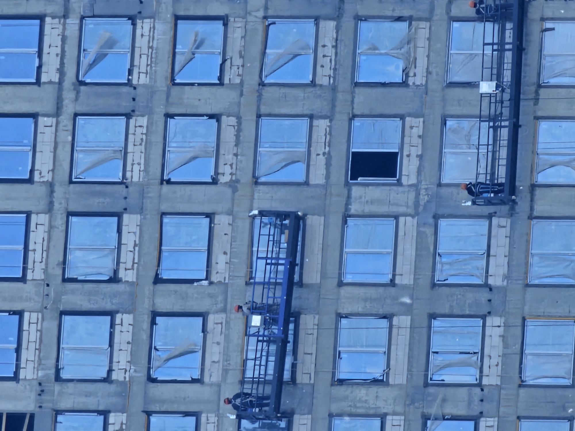 The image displays the facade of a building undergoing construction or renovation in Moscow, Russia. The structure is characterized by a repetitive grid of rectangular windows set within a grey concrete-like exterior. Around many of the window frames, exposed lighter-colored, possibly brick or block-like, material is visible. Most windows feature a reflective, light blue surface, which appears to be a protective film, with several showing torn or partially detached sections. One window on the fourth row from the top, second from the right, is open, revealing a dark interior.
Two blue vertical lifting platforms, likely scaffolding or mast climbing work platforms, are attached to the building's exterior. One is positioned on the right side, and another towards the left-center. Two construction workers, each wearing an orange hard hat and dark work clothing, are visible on the lower sections of these platforms. They appear to be actively engaged in work on the building's facade or operating the machinery. The scene depicts a daytime setting under clear weather conditions, focusing on the ongoing work at various levels of the building. There is no discernible text visible in the image.