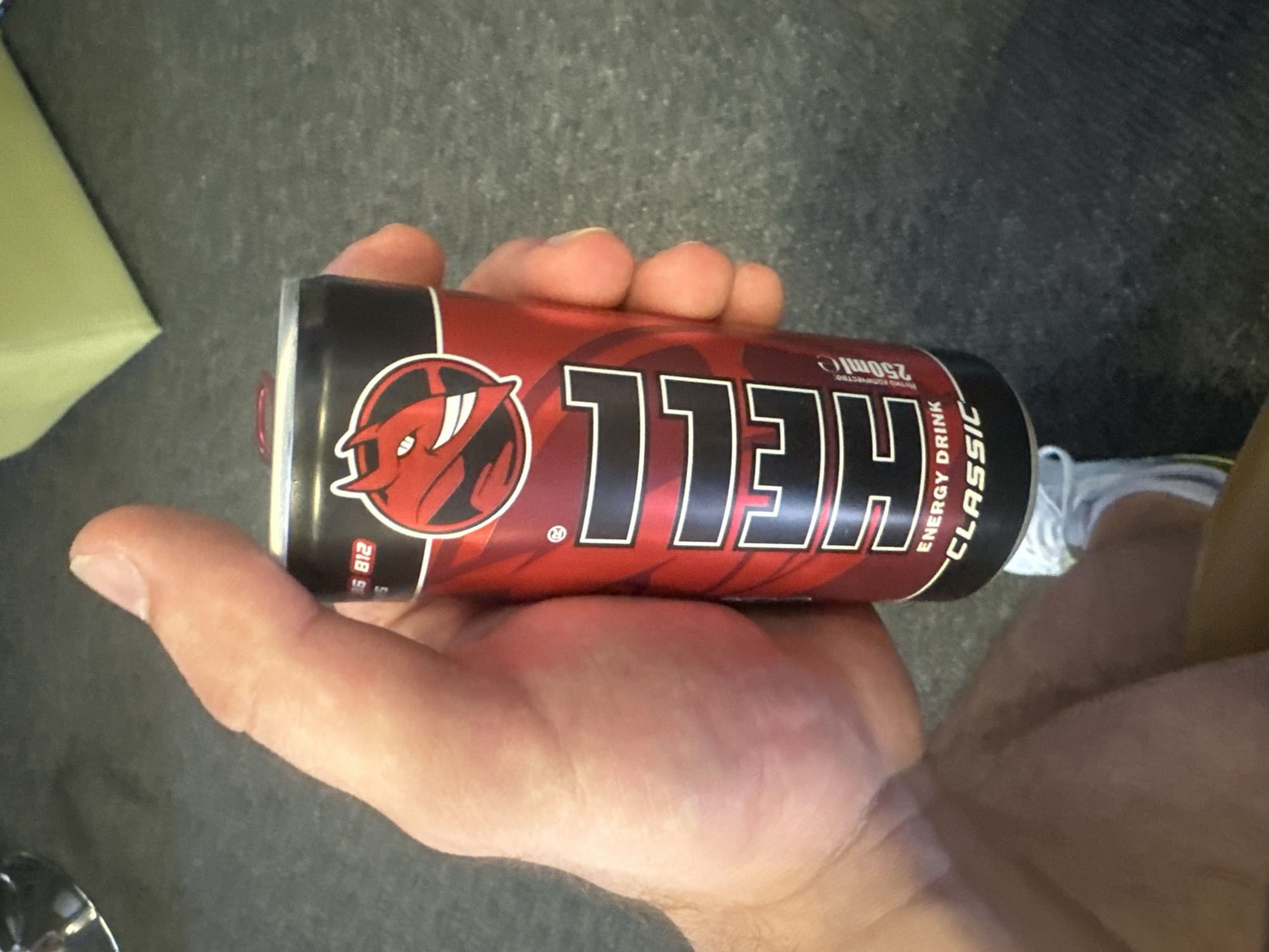 The image displays a close-up, angled shot of a human hand holding a red and black "HELL Classic Energy Drink" can. The hand, likely an adult's, grasps the can, with fingers visible over the top and the thumb resting on the side. The can is predominantly red with a black section, featuring a bold, black-outlined "HELL" logo and a distinct red devil/demon head emblem to its left. Vertical white text on the can clearly states "ENERGY DRINK" and "CLASSIC," alongside "250ml e." Smaller text, possibly in Hungarian, such as "RETURNA KÚPUNKBAN" and "Minden jog fenntartva," and a registered trademark symbol are also discernible. Red numbers "218 9" are visible on a black band near the can's base.
The background is a blurred, dark, and textured surface, possibly concrete or a dark floor. Faint hints of a light-colored object or fabric appear in the upper left, and a reflective surface is visible in the lower left. A partial view of a white shoe or sneaker can be seen in the bottom right corner. The lighting is ambient, suggesting either an indoor environment or an outdoor setting with indirect light. The scene captures the simple act of a person holding the energy drink. The provided location context for this image is Blagoevgrad, Bulgaria.