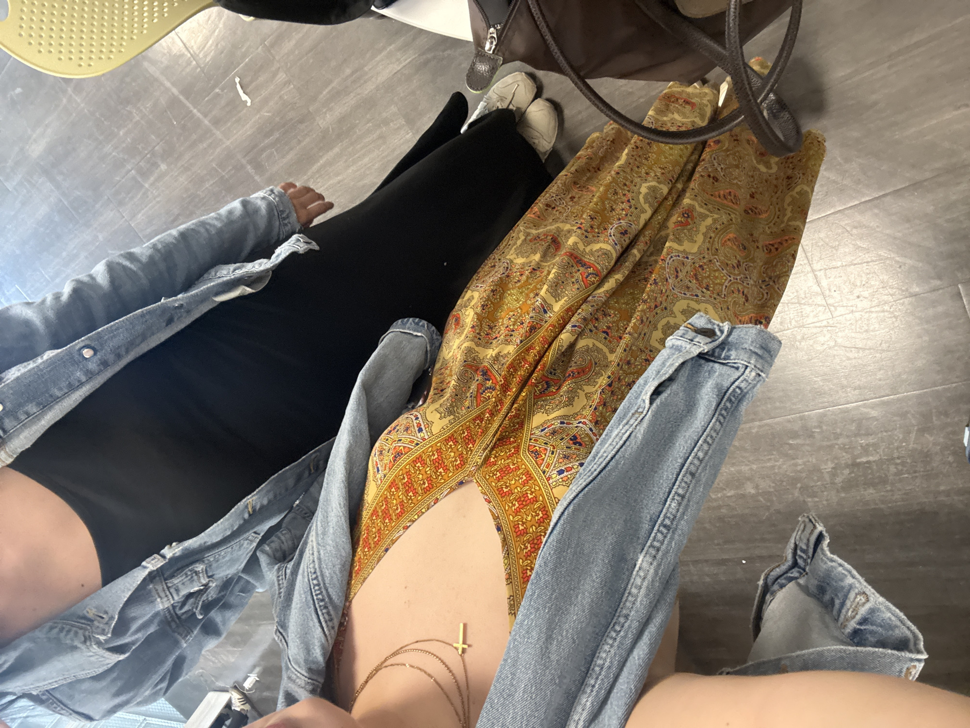 An overhead shot captures two individuals seated or lounging indoors on a light grey, possibly tiled or concrete floor. The perspective looks down upon their torsos and legs.
On the left, one person wears a black top or dress, covered by a light blue denim jacket with visible wear and tear. Their arm is bent, and a hand is visible. Below, their legs are covered in black fabric, and white sneakers are partially visible near the top of the frame.
On the right, another person wears a garment featuring a vibrant yellow and orange paisley pattern, possibly a dress or a wide-legged jumpsuit, also paired with a light blue denim jacket. This person’s bare midriff is visible, adorned with layered golden necklaces, including one prominent inverted cross pendant. Their legs are partially visible, covered by the patterned fabric.
Other elements include a partial view of a light yellow, perforated object (possibly a chair or tray) in the upper left, and a dark brown bag or strap near the top center. The overall setting appears casual and indoor, with no specific activities or explicit location markers visible within the frame. There is no discernible text in the image. The time of day and specific emotions are not evident.