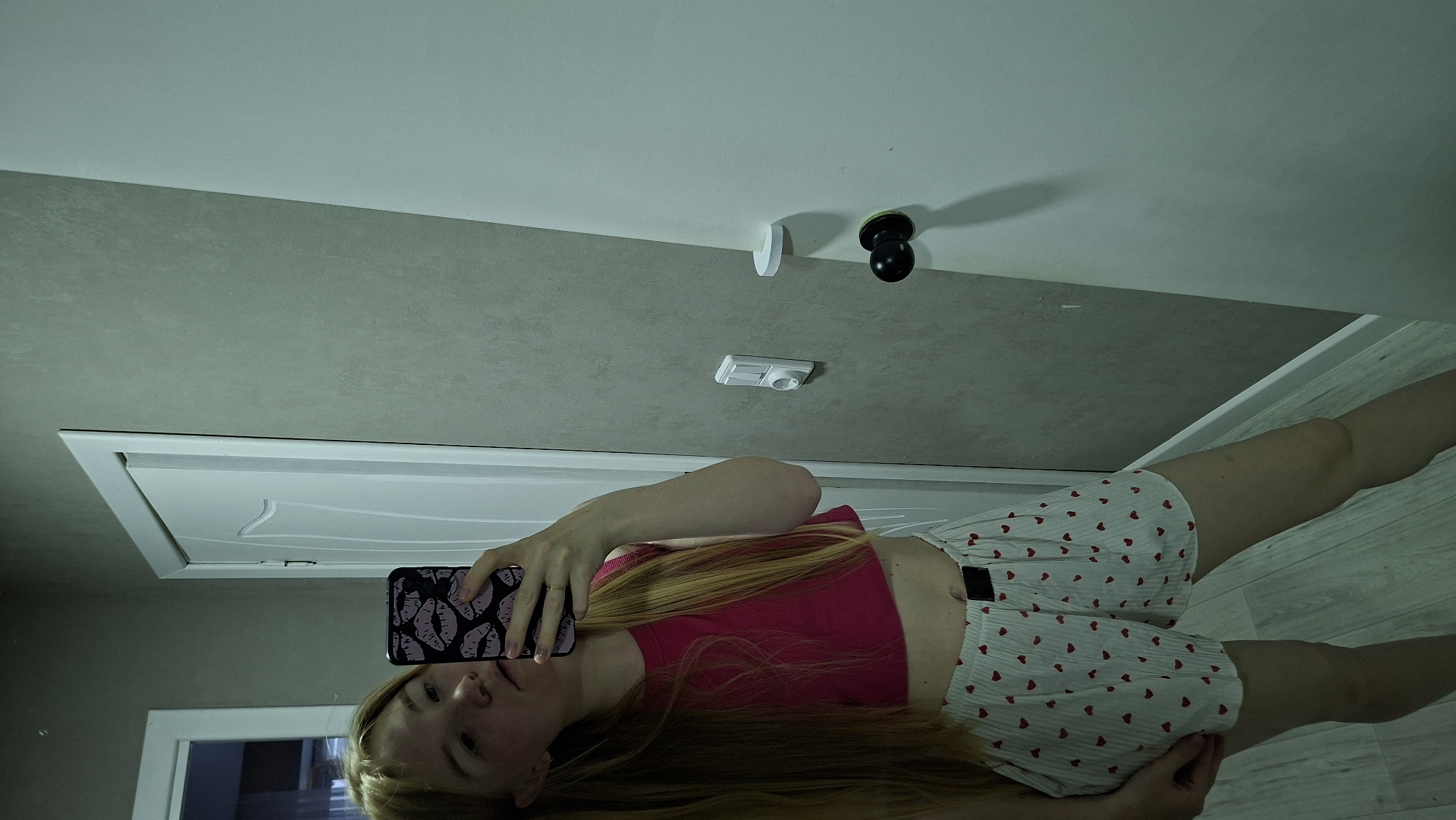 A young woman with long blonde hair is taking a mirror selfie in what appears to be a residential hallway. She is wearing a bright pink cropped tank top and white shorts patterned with small red hearts. Her phone, with a phone case decorated with lips, is held up to capture the image. The background shows a textured gray wall with a white door on the right side of the frame, featuring a black doorknob and a white door handle. To the left, another white door is visible, partially open, revealing a glimpse of another room. The lighting is dim, casting shadows on the walls. This scene is consistent with an indoor setting within a private residence in Chaikovsky, Russia, given the user's request for location context.