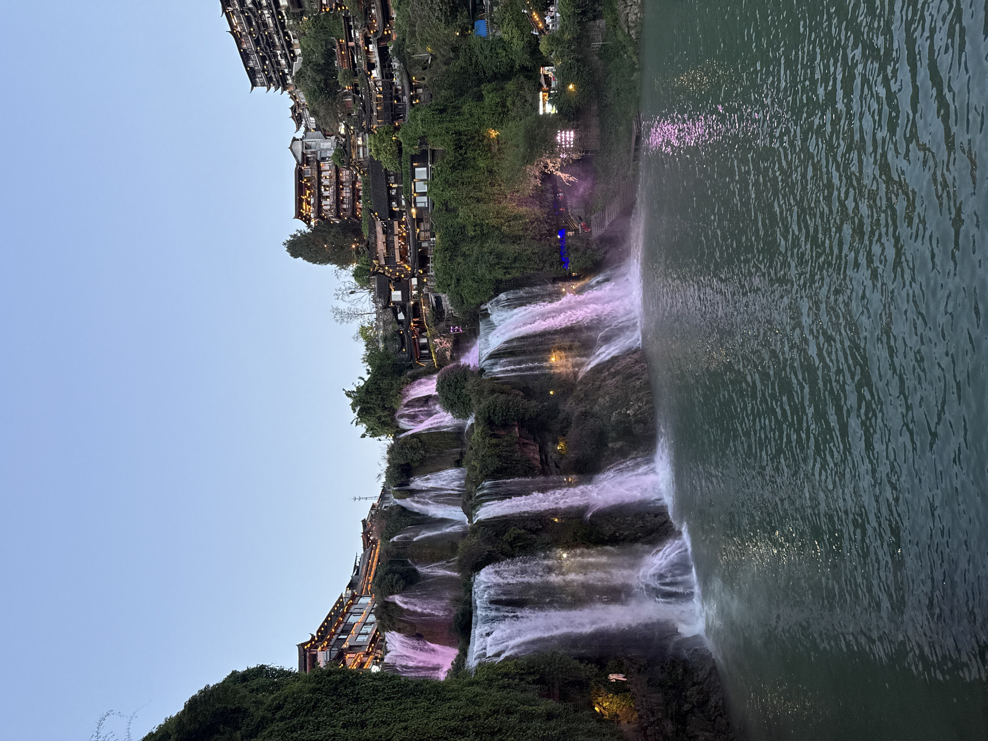 The image shows the illuminated Furong Waterfall in Furong Ancient Town, Hunan Province, China. Water cascades down a tiered rock formation, creating a series of waterfalls. Vegetation grows along the cliffs, intertwined with buildings perched on the edges. The water reflects pink and white light from the artificial lighting used to illuminate the scene. The sky is light blue. The water flows into a body of water that appears dark in color.