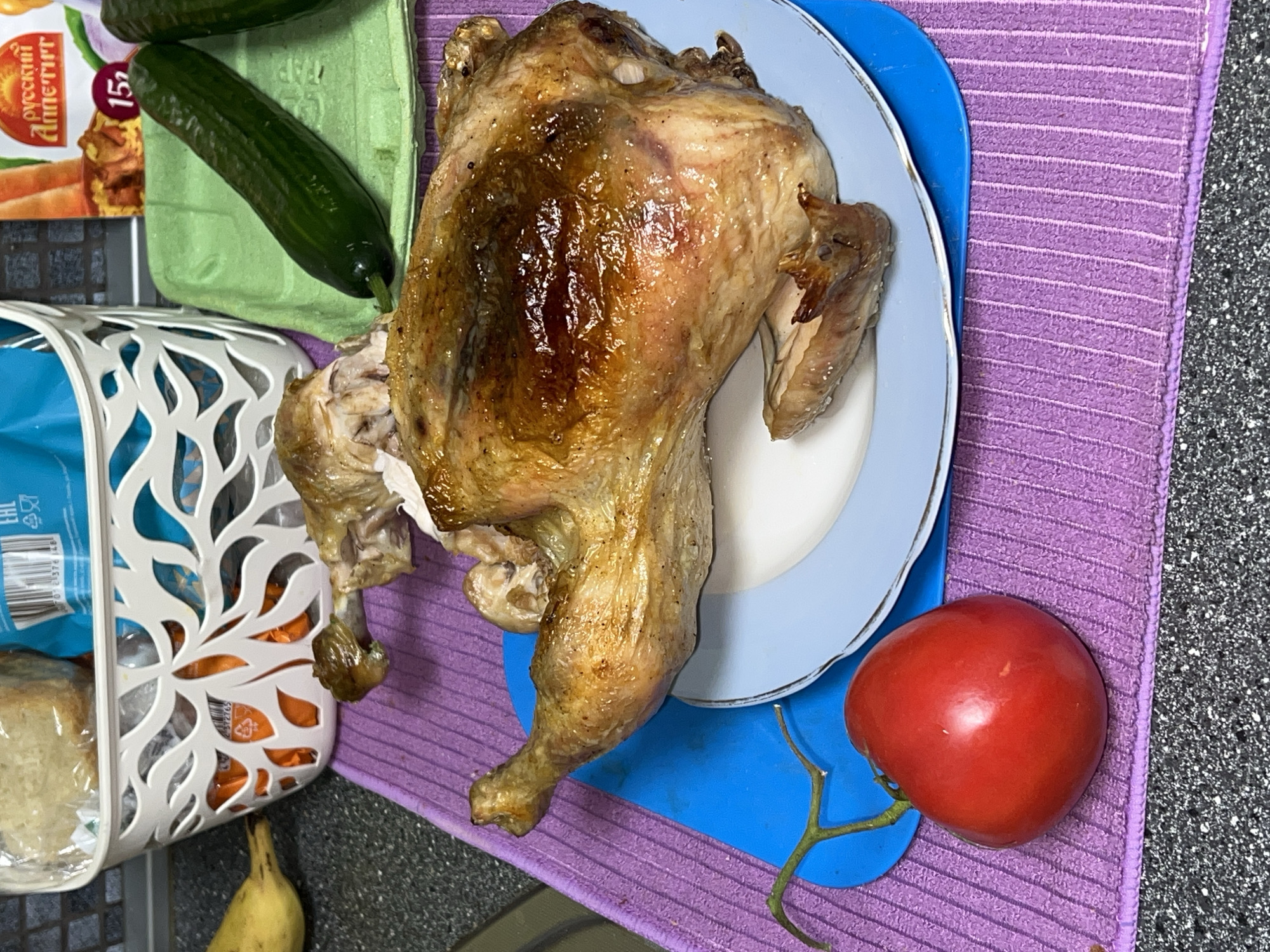 This image captures a casual home meal setting, likely in a kitchen or dining area, with various food items arranged on a dark grey, speckled countertop.
The central subject is a whole roasted chicken, golden-brown and glistening, presented on a light blue plate with a white rim. The chicken appears recently cooked, with one leg partially torn or detached. The plate rests on a blue plastic cutting board, which in turn is placed on a light purple, ribbed mat.
To the left of the chicken, two green cucumbers are visible. One lies on a light green egg carton, while the other is partially obscured behind it. Further to the left, a white decorative plastic basket holds what appears to be sliced bread wrapped in plastic, and possibly other items like orange packaging. A blue package with the "EAC" conformity mark is also visible inside the basket.
A single vibrant red tomato, still attached to a small portion of its vine, rests on the blue cutting board next to the chicken. At the very bottom of the frame, partially visible, is a yellow banana or banana peel.
A food package with the text "РУССКИЙ АППЕТИТ" (Russian Appetite) and "15г" (15g) is seen in the upper left, suggesting the context of Perm, Russia. The scene depicts a simple, prepared meal, possibly for a family. No people are visible, and the lighting is typical for an indoor setting.
