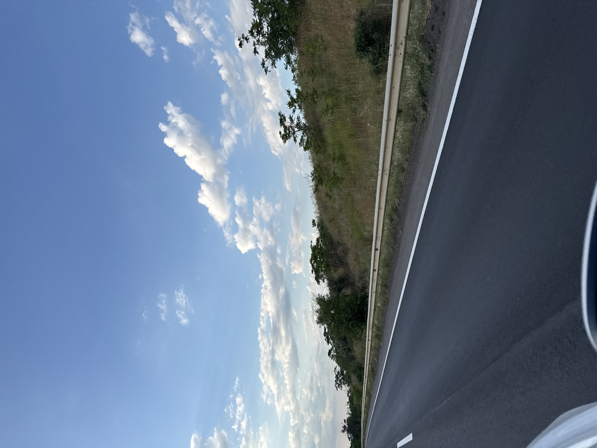 A view from a moving vehicle on a highway near Altestove, Ukraine shows an asphalt road with a white lane divider and a metal guardrail. The road runs diagonally across the frame from the lower left to the upper right. Beyond the guardrail, a grassy embankment rises, dotted with sparse green trees and dry vegetation. The sky above is a clear blue with scattered white cumulus clouds. The image captures a typical roadside scene on a sunny day.