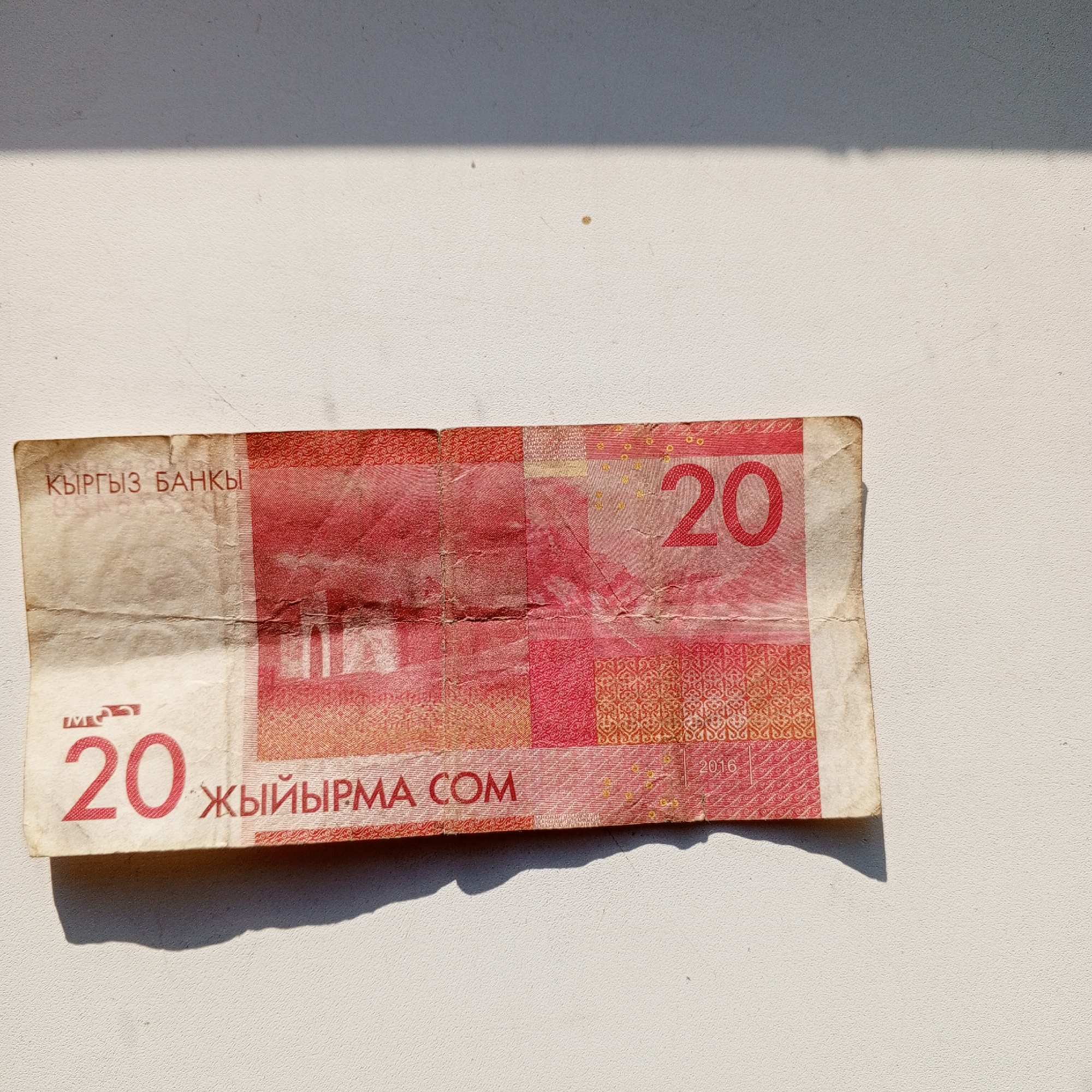 The image shows a wrinkled, red and orange twenty som banknote, with "КЫРГЫЗ БАНКЫ" printed in red at the top left and "20" printed in large red font on the left side. The word "ЖЫЙЫРМА" is printed in red to the right of the "20" on the bottom. The year "2016" is visible in the top right corner. The banknote is lying on a textured, light-colored surface, with a shadow cast across it from an unseen object. The location is Sayanogorsk, Russia.