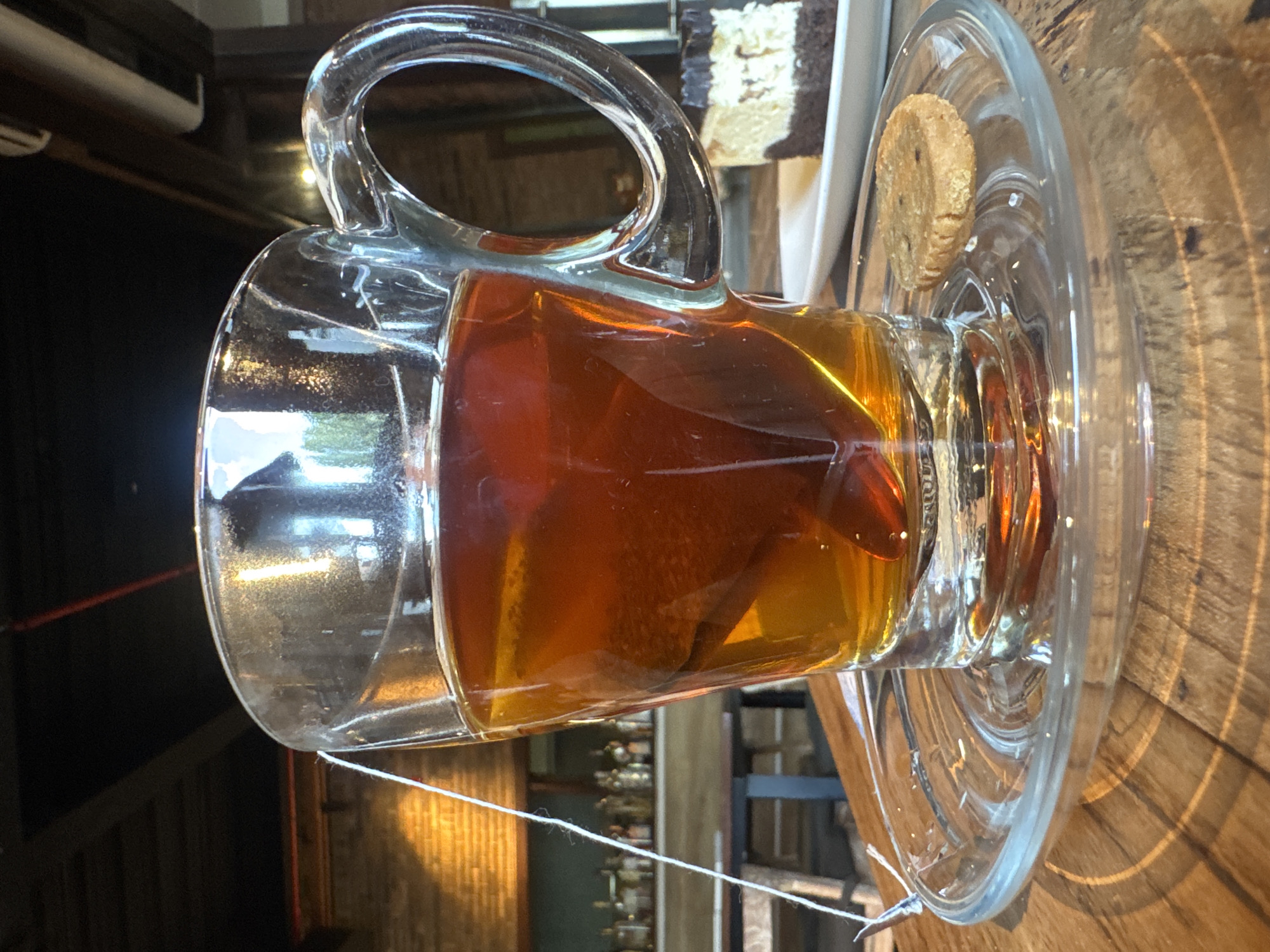 A close-up shot features a glass mug filled with amber-colored tea, complete with a tea bag submerged within. The mug rests on a clear glass saucer, which in turn sits on a wooden table. A single cookie is placed on the saucer to the right of the mug. In the background, out of focus, a slice of chocolate and cream cake is visible on a white plate. The setting appears to be indoors, possibly a cafe or restaurant, with wooden beams and brick walls suggested in the blurred backdrop. The overall ambiance is warm and inviting, typical of a relaxing cafe environment in Kuta Selatan, Indonesia.
