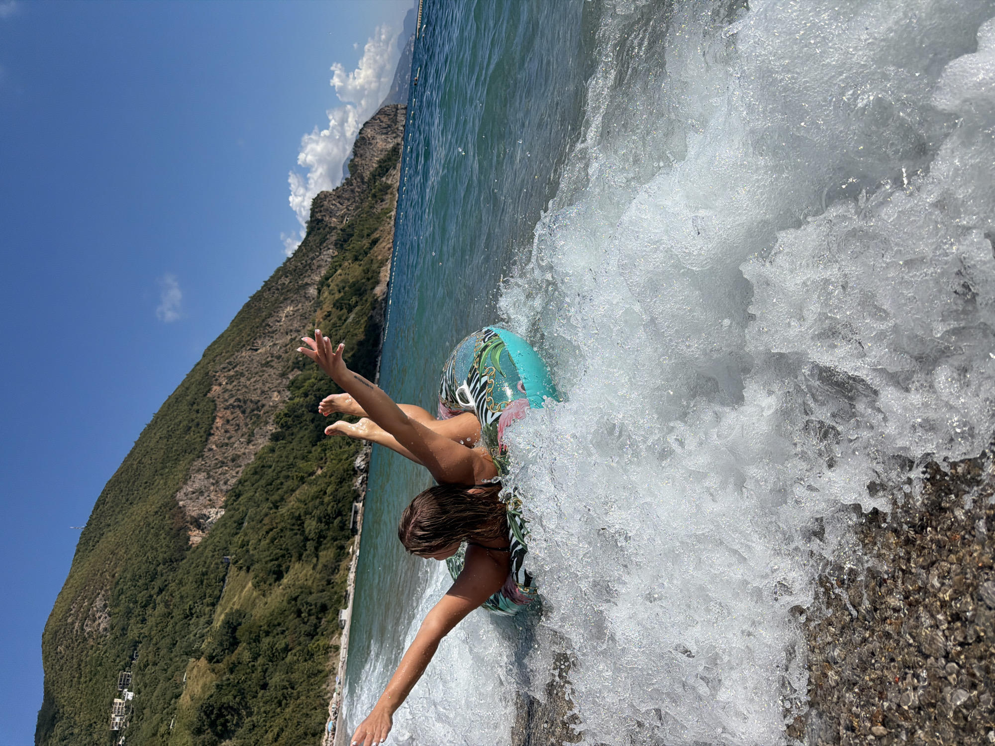 A young woman, partially visible, floats on her back in a turquoise inflatable ring with a tropical pattern. She is in shallow, breaking waves near a pebble beach. Her arms are outstretched and her legs are raised out of the water, creating splashes. The background shows a steep, tree-covered mountainside, characteristic of the area near Prijevor, Montenegro. The sea is a vibrant blue, and the sky is clear with a few scattered clouds. The overall scene depicts a moment of playful enjoyment on a sunny day at the coast.