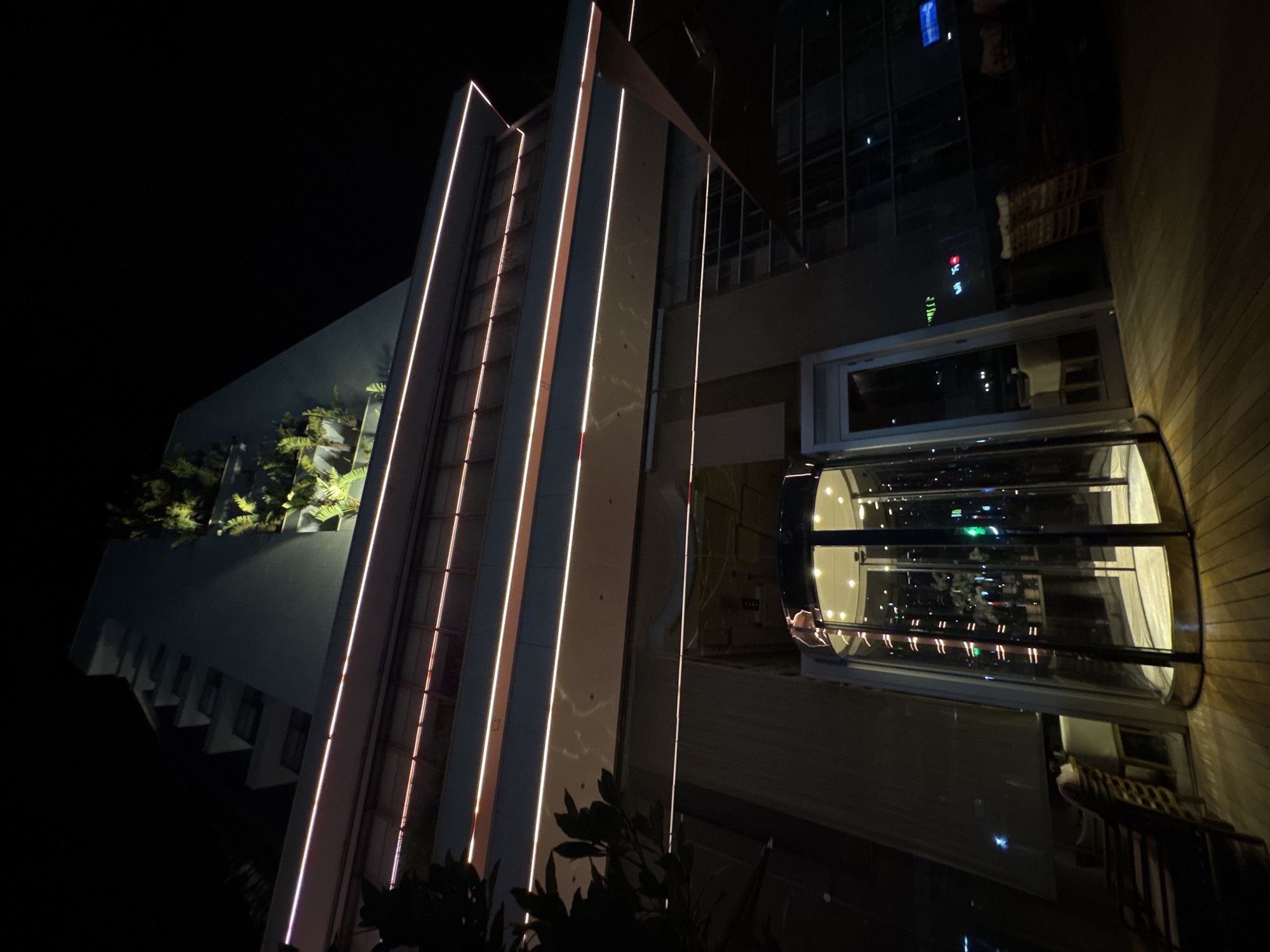 A low-angle, night-time shot captures the exterior of a modern, multi-story building in Agios Tychona Community, Cyprus. The building features a stepped facade with lush greenery cascading down one side. Horizontal lines of white and pinkish LED lights accentuate the building's architecture across multiple levels.
In the foreground, a revolving glass door entrance is illuminated from within, showcasing what appears to be a reception area or lobby. Beyond the entrance, to the left, is a seating area with outdoor furniture. To the right, there's a wooden deck with more seating, partially obscured by an awning. The overall scene is dark, with artificial lights from the building and the entrance providing illumination.