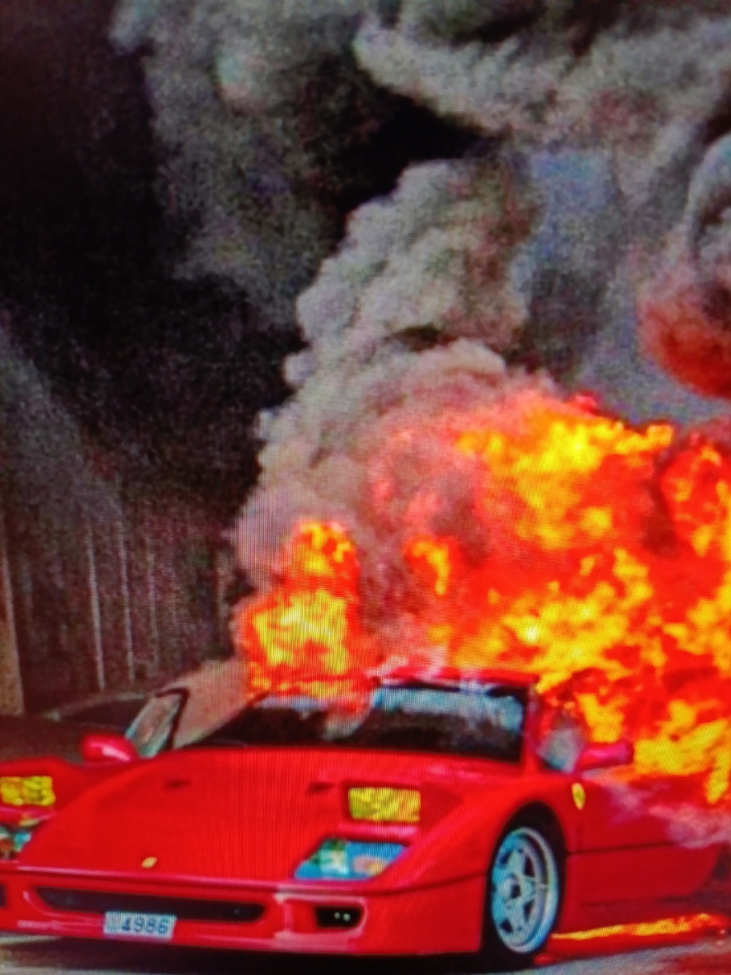 In an urban environment, a red Ferrari F40 is engulfed in flames. The vehicle is positioned on what appears to be a street or roadway. Intense orange and yellow flames erupt from the engine compartment and spread across the rear of the car. Thick, grey smoke billows upwards and outwards, obscuring the background, which consists of dark, indistinct structures that may be buildings or fencing. The car's front is visible, showing its distinct headlights, front bumper, and a license plate that reads "4986". The car's wheels are also visible, with the rear wheel on the right side of the image displaying a silver alloy rim. No people are visible in the scene. The overall impression is of a dramatic and destructive event involving the burning vehicle.