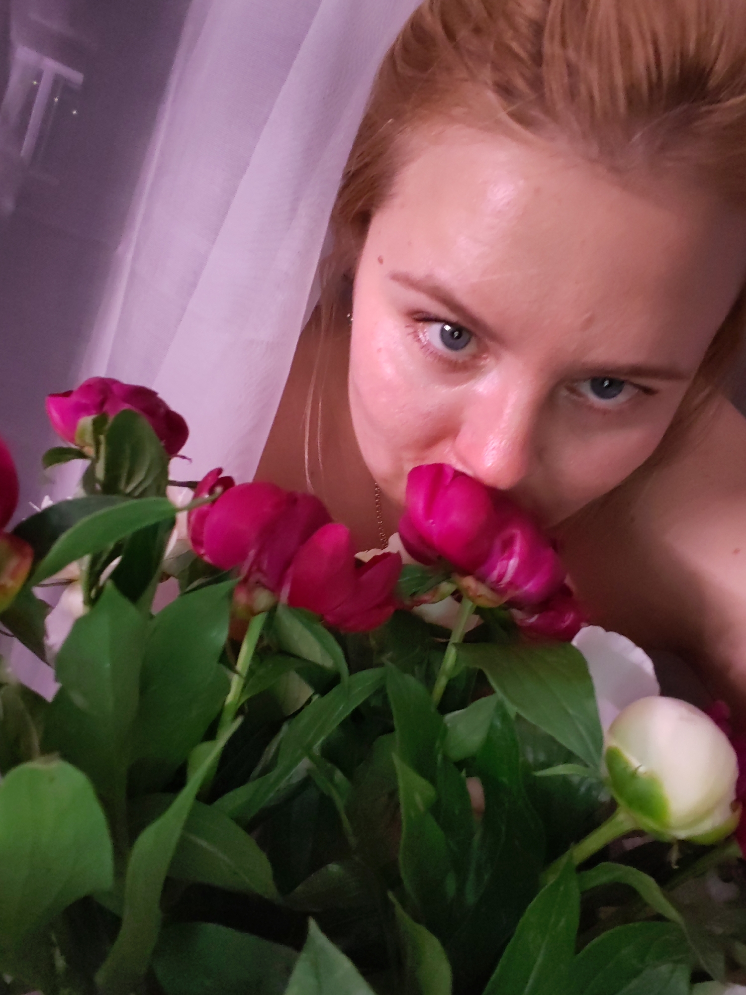 A young woman with blonde hair is shown in a close-up shot, leaning forward to smell a bouquet of pink and white peonies. She has blue eyes and fair skin. The background is a soft, blurred pastel color, possibly a curtain or sheer fabric, with a hint of window light. The image is captured in a dimly lit indoor setting. There is no indication of the location being in Izhevsk, Russia, or any specific city.