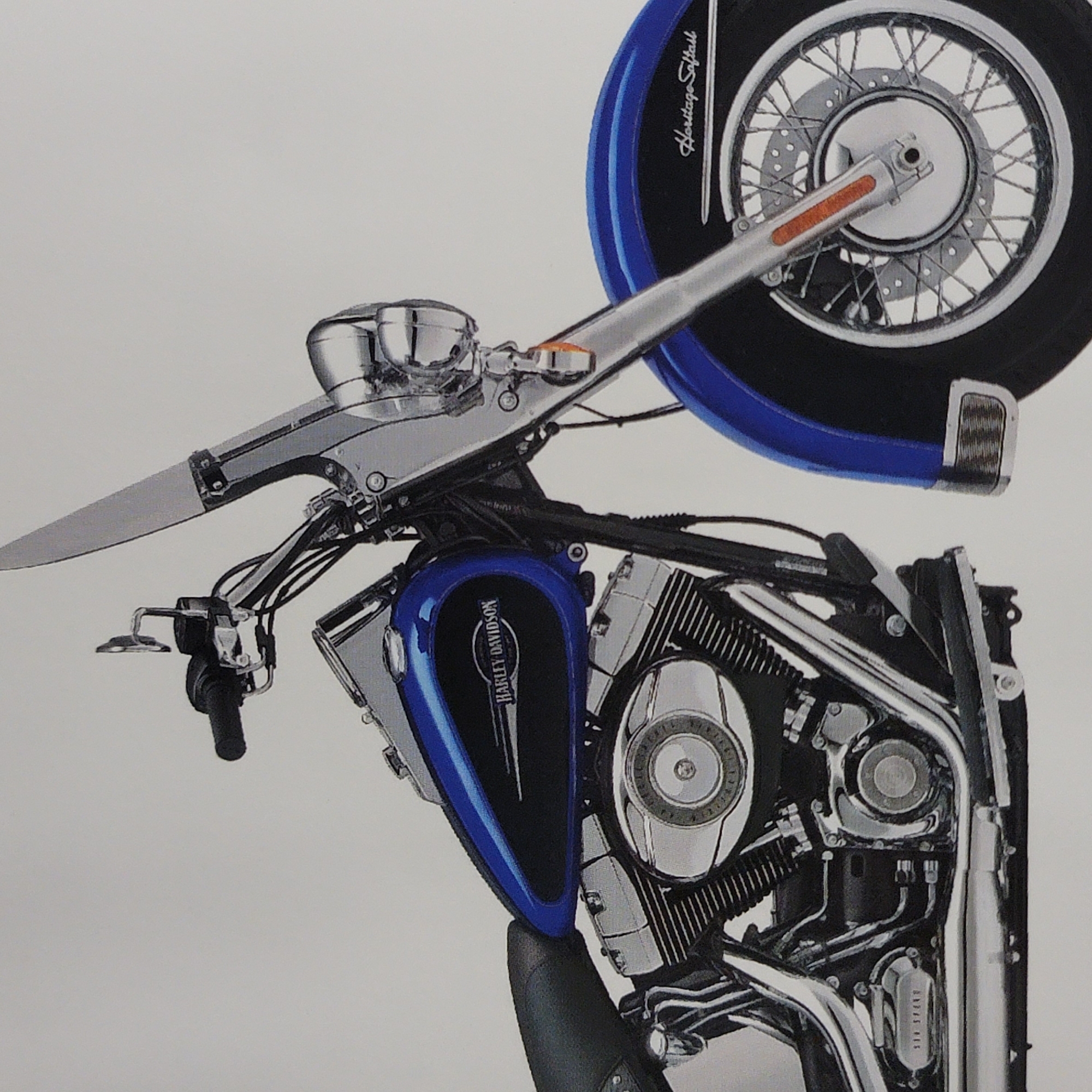 A side profile of a blue and silver Harley Davidson motorcycle is captured against a plain white background. The motorcycle features a prominent windshield, chrome accents on the engine and handlebars, and a black leather seat. The "Harley Davidson" logo is visible on the fuel tank, along with "Heritage Softail" text on the front fender. The motorcycle is stationary and appears to be a studio shot or a promotional image, as there is no surrounding environment or indication of a city location.