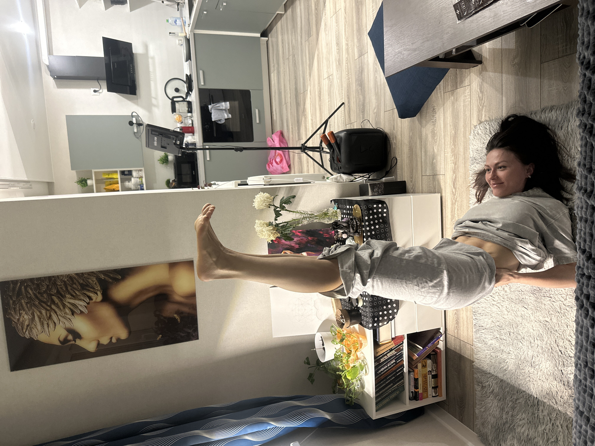 Here is a detailed description of the image:
**Scene:** The image captures the interior of a modern apartment, likely in a residential area. The location may be in Murino, Russia, although this is not directly verifiable from the image itself.
**People:** A woman is featured prominently in the foreground. She is lying on a rug, wearing casual gray clothing. Her dark hair is loose around her face, and she is smiling slightly. Her legs are elevated.
**Objects:**
* **Furniture:** There is a table with a blue and grey surface near the woman. A white shelving unit houses books and plants.
* **Decor:** A painting or printed artwork featuring a classic headshot is mounted on the wall. Floral arrangements are on the shelving unit.
* **Electronics:** A television and sound system are present.
* **Kitchen:** Kitchen appliances and cabinetry are visible.
**Actions/Interactions:** The woman is relaxed, seemingly resting in the living space. The scene appears staged.
**Environment:** The apartment has light-colored walls and wooden floors. The lighting is bright, and the space feels clean and organized. The scene seems modern and recently built.